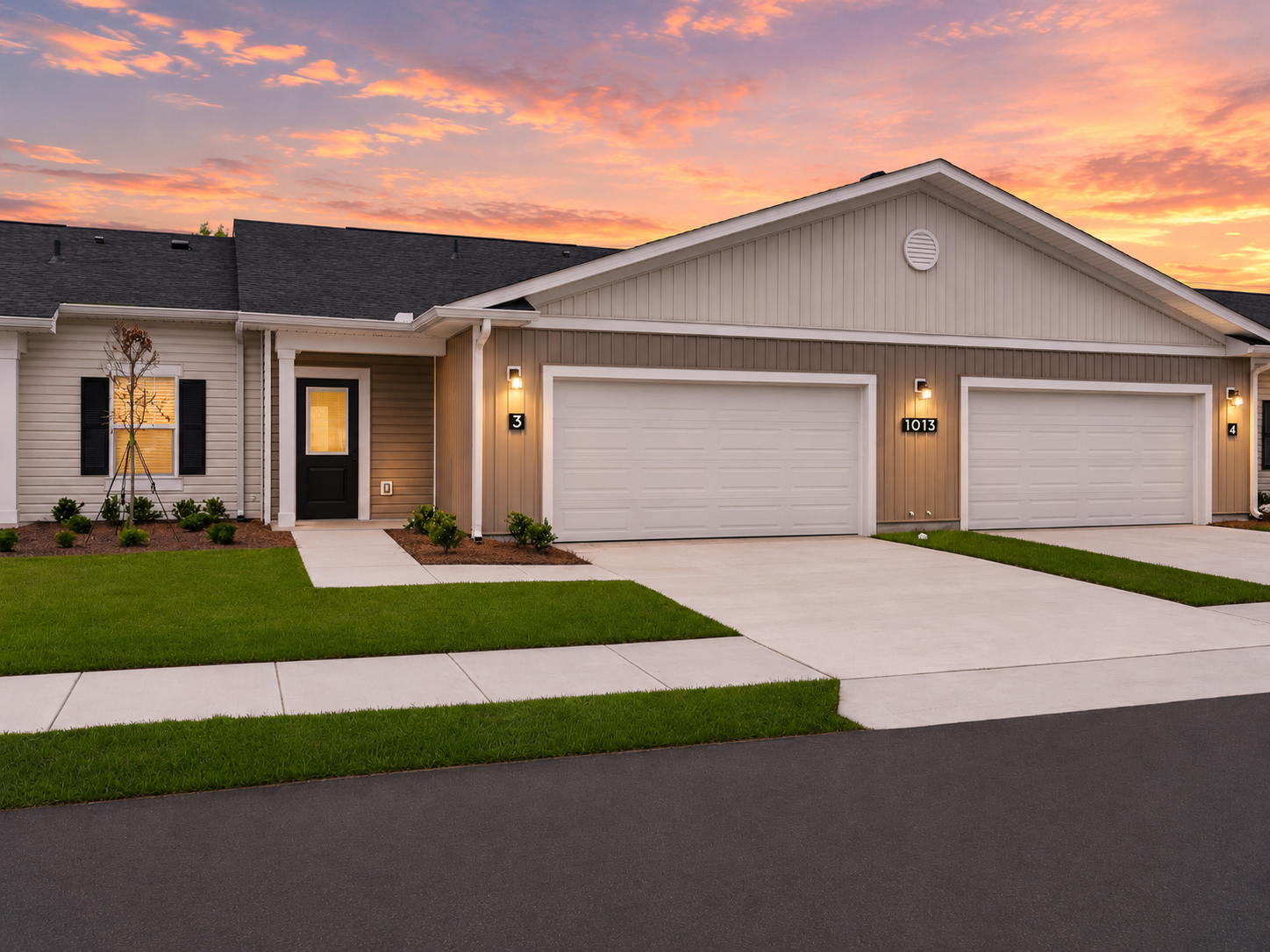 A modern residential building featuring a two-car garage and a well-maintained lawn. The exterior is a blend of light and dark siding with a welcoming front door. The setting is during sunset, with a colorful sky in the background, enhancing the home's appeal.