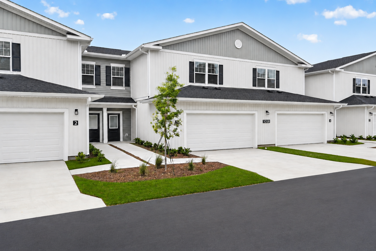 A modern residential building with a gray and white exterior, featuring multiple garages and a neatly landscaped front yard. The driveway is paved, and there are small trees and plants along the path leading to the entrance. The sky is clear with a few clouds.