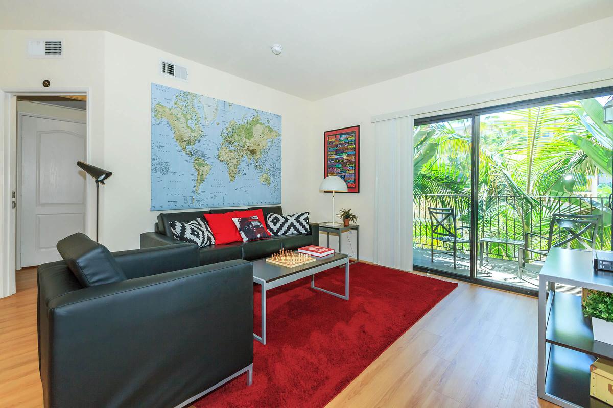 A cozy living room featuring a black leather sofa with red and black cushions, a glass coffee table with a chess set, and a large world map on the wall. There are large windows overlooking a patio with greenery, and the room is decorated with a red area rug and various wall art.