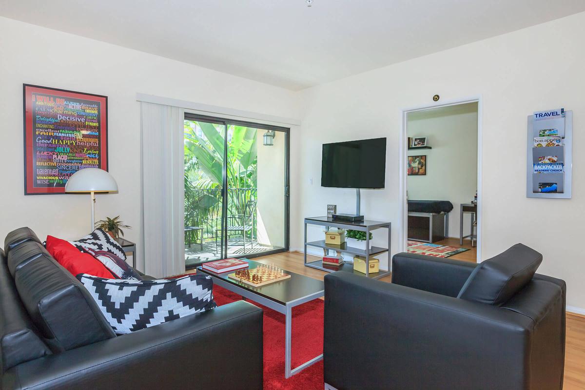 A modern living room featuring black leather sofas with red and white accents, a coffee table on a red rug, and a television mounted on the wall. There are sliding glass doors leading to a balcony with greenery visible outside. Wall decor includes a colorful framed poster and a travel-themed display on the opposite wall.