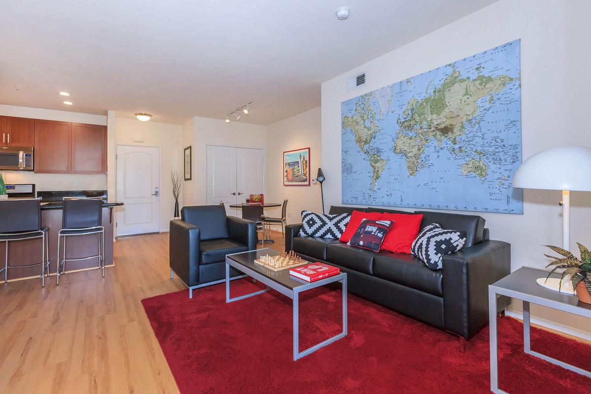 A stylish living room featuring a black sofa with red and white cushions, a red area rug, and a world map on the wall. There is a coffee table with a chess set and decorative items, as well as a small dining area in the background. The kitchen is visible with modern cabinetry and appliances.