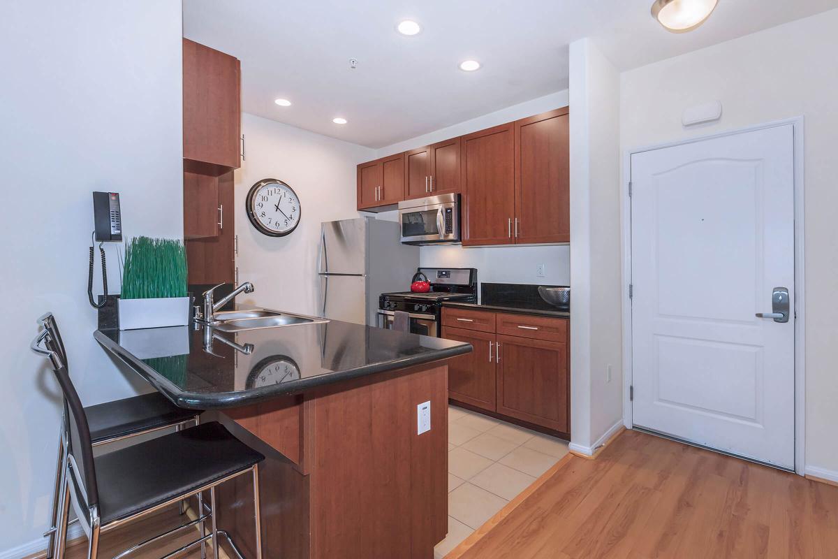 A modern kitchen featuring dark wooden cabinets, a stainless steel refrigerator, an oven, and a sink with a black granite countertop. There's a round wall clock on the wall, under-cabinet lighting, and a stylish bar stool at the counter. The floor has light-colored tiles and there is a door leading to another room.