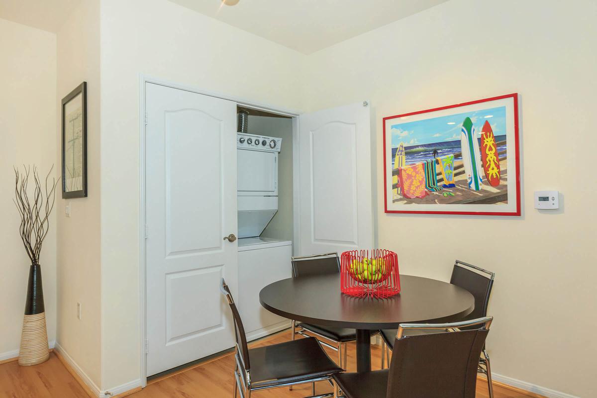 A small dining area featuring a round table with black chairs, a colorful beach-themed artwork on the wall, and a laundry closet with doors partially open, revealing a stacked washer and dryer. A decorative fruit bowl sits at the center of the table, and there is a tall plant in a vase in one corner.