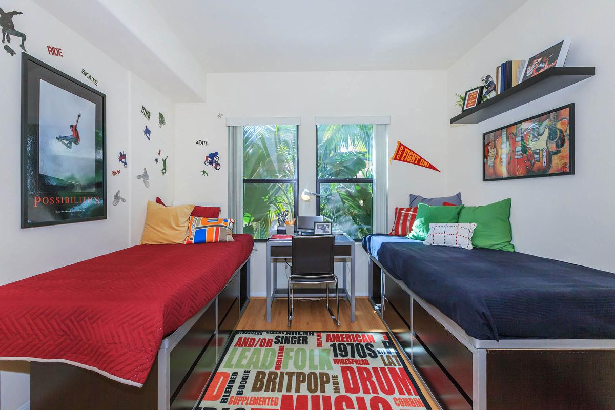 A bright room with two twin beds, one with red bedding and the other with navy blue. There's a small desk with a black chair between the beds. The walls are decorated with posters and decals related to sports, and there are two windows letting in natural light, revealing greenery outside. A colorful area rug features various text designs.