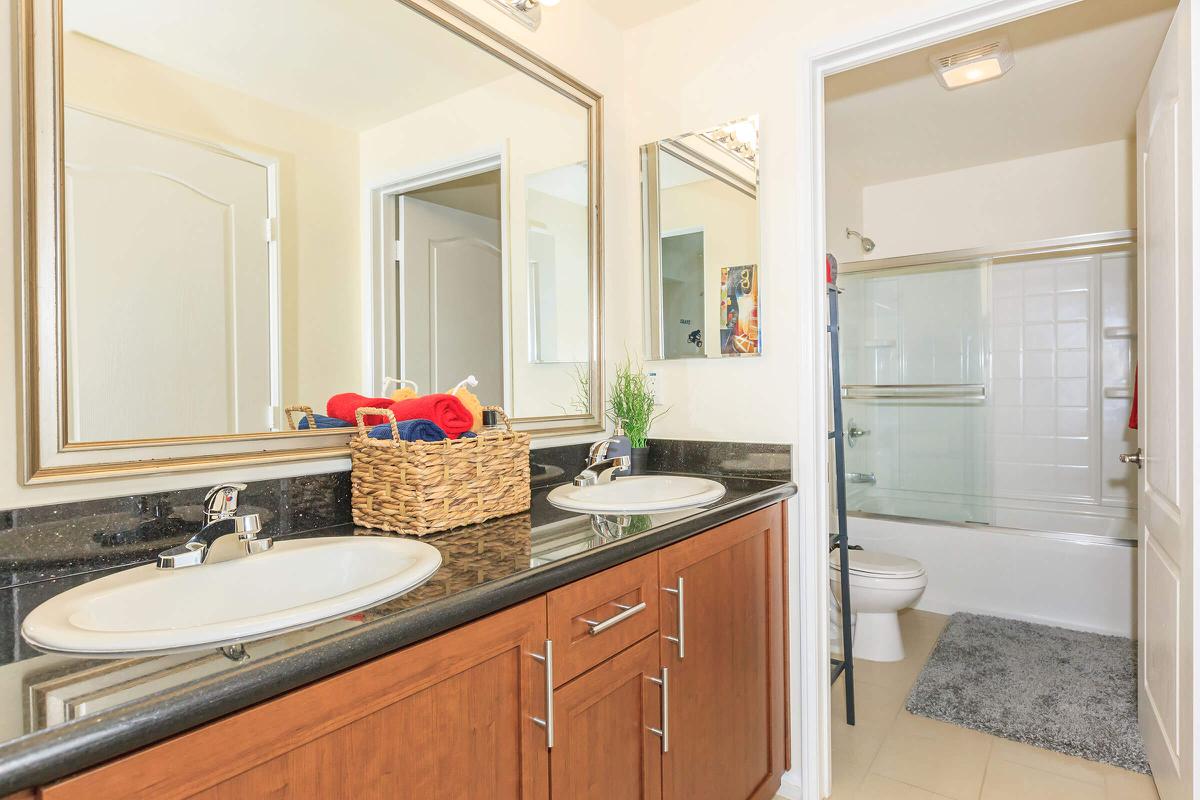 A modern bathroom featuring a double vanity with two sinks, mirrors above each sink, a dark granite countertop, and wooden cabinetry. A woven basket contains towels, and there is a shower/tub combination visible in the back. The flooring is light tile, and a small rug is placed in front of the sinks.