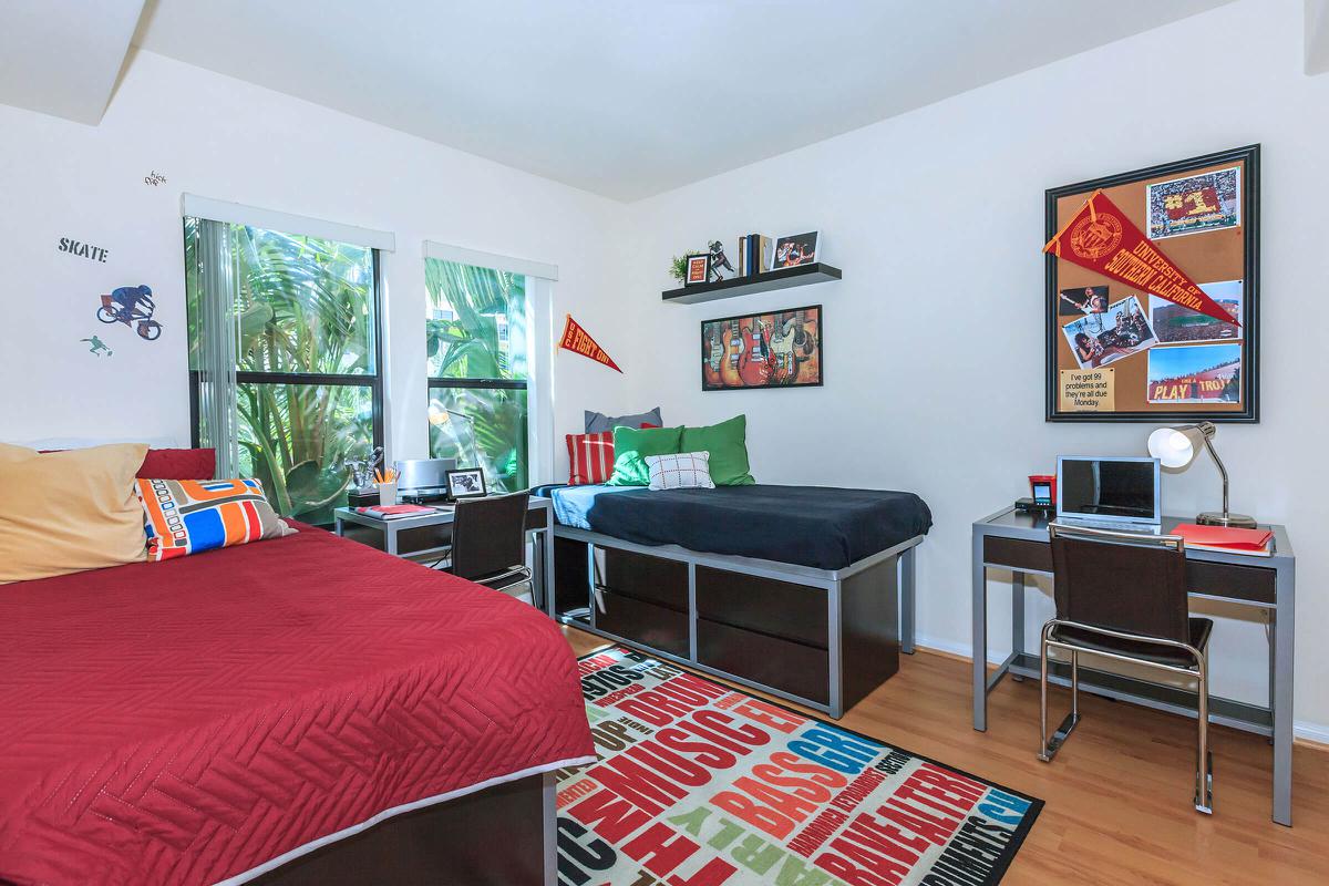 A brightly decorated shared bedroom featuring two beds, one with a red comforter and the other with a dark blue blanket. The room has large windows with greenery outside and is adorned with a variety of decorative pillows. There are shelves on the wall displaying memorabilia, along with a bulletin board. A desk with a laptop and chair is positioned next to the bed, and a colorful area rug covers the floor.