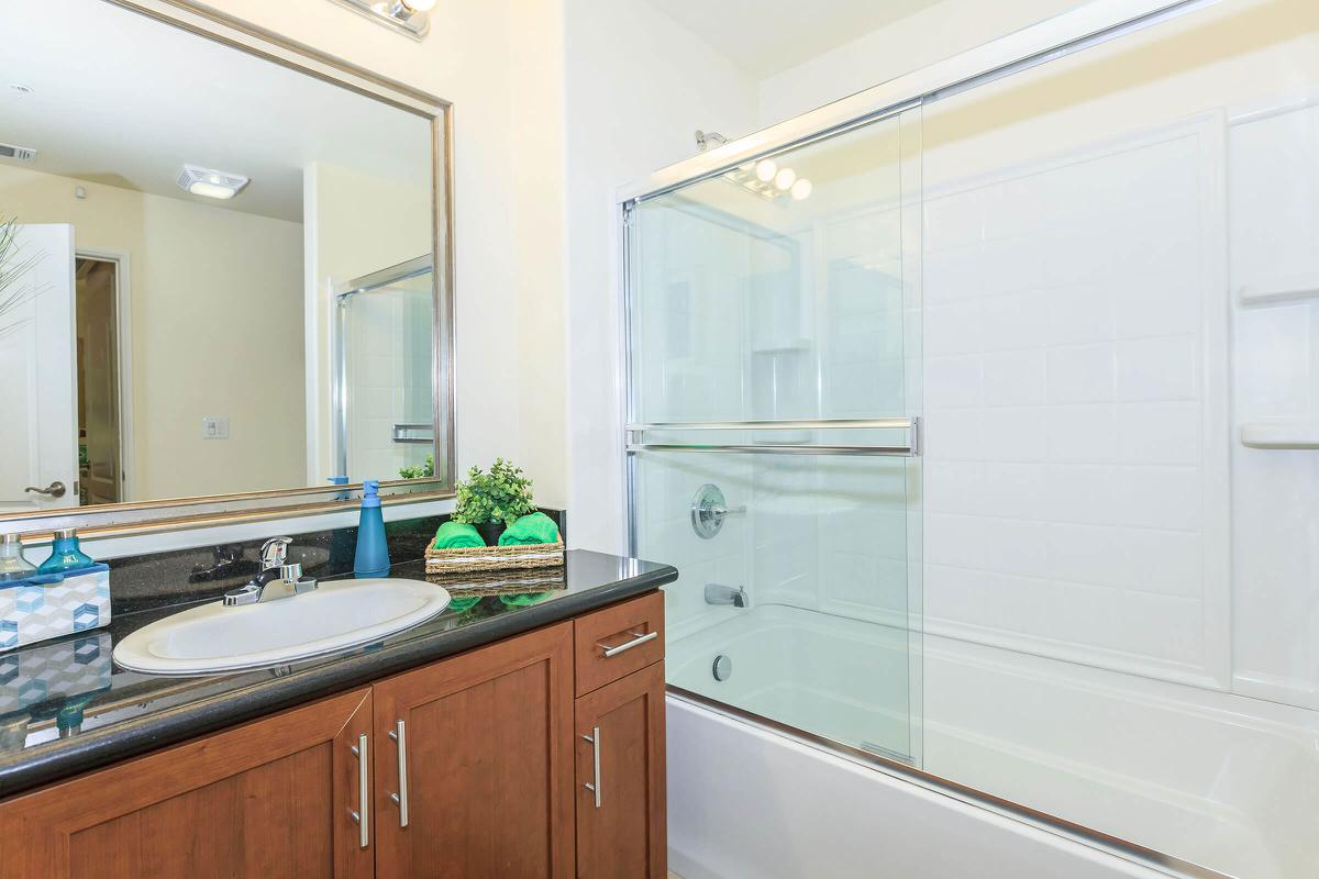 A modern bathroom featuring a bathtub with a glass shower enclosure, a sink with a dark countertop, and a large mirror. Decorative items include a small basket and blue accents on the sink. The walls are light-colored, and there is adequate natural light.