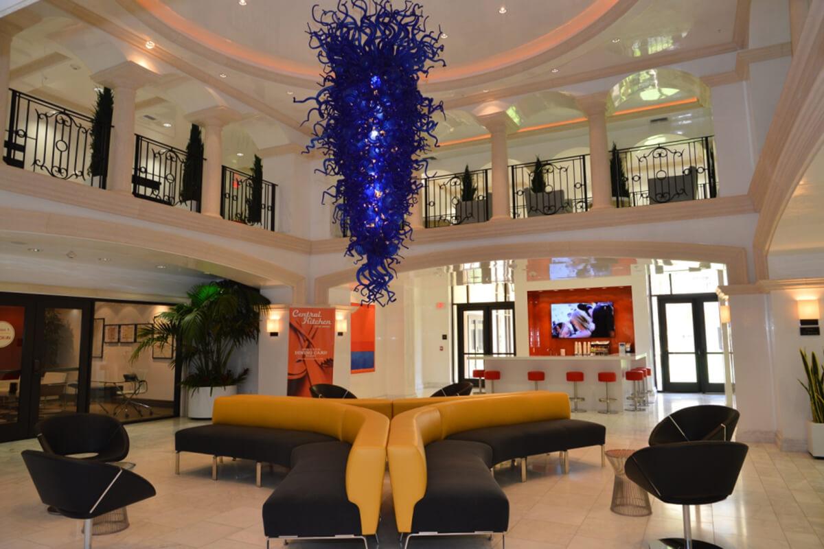 A modern lobby featuring a large, abstract blue chandelier hanging from the ceiling. The space has a high ceiling with elegant arches, a reception area with contemporary seating arrangements in yellow and black, and plants adding greenery to the environment. Large windows allow natural light to fill the space.