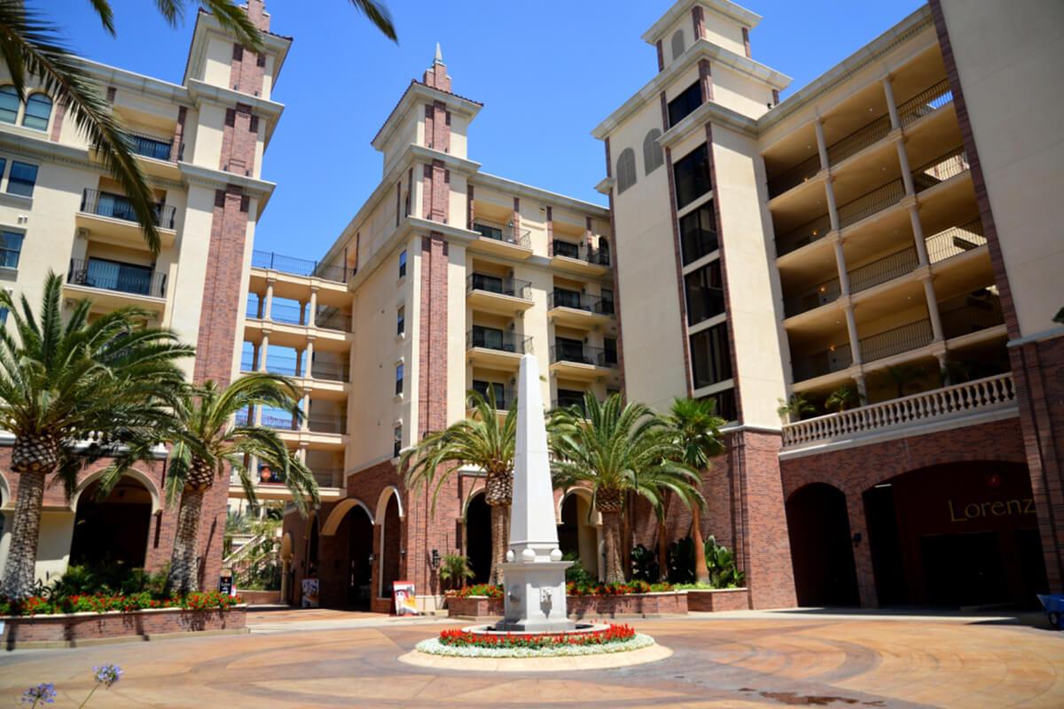 A sunny courtyard featuring a central fountain surrounded by palm trees and tall buildings with balconies. Decorative landscaping includes colorful flowers and a pathway leading through the area.