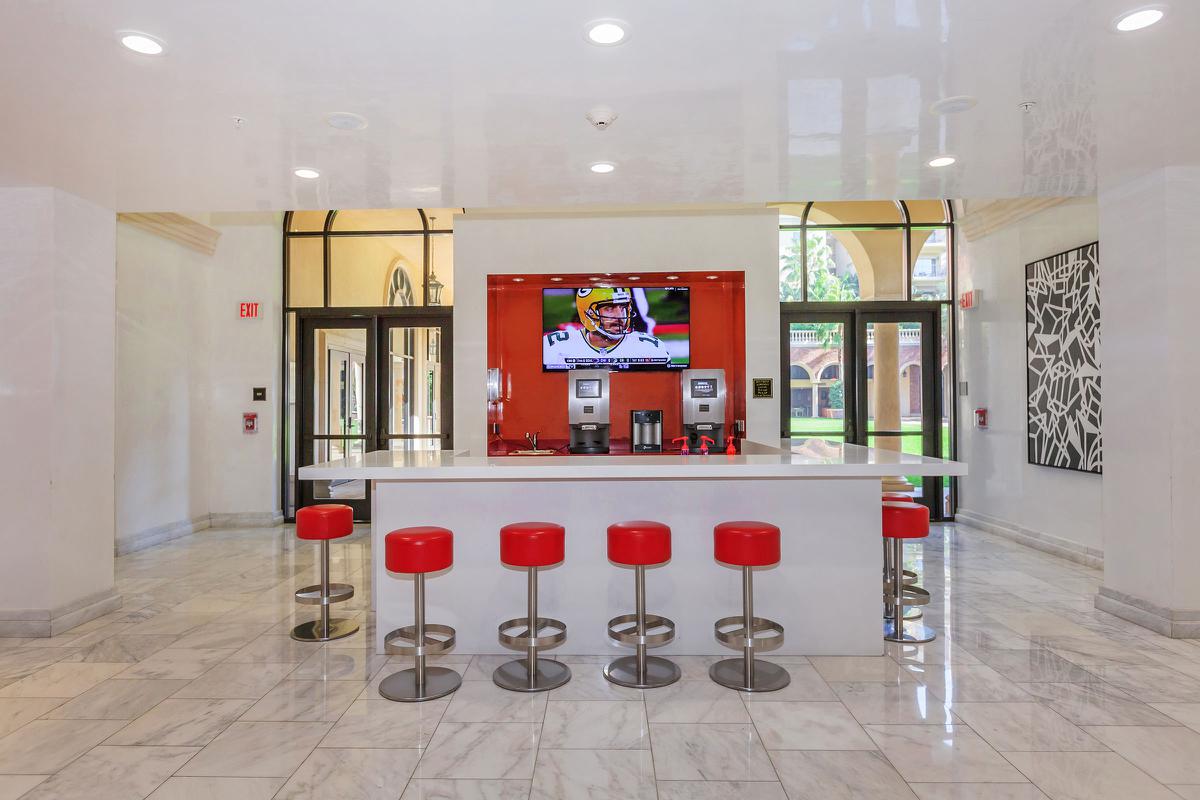 A modern reception area featuring a white countertop with red stools, a large television screen displaying a sports game, and an open, airy design with marble flooring and glass doors.