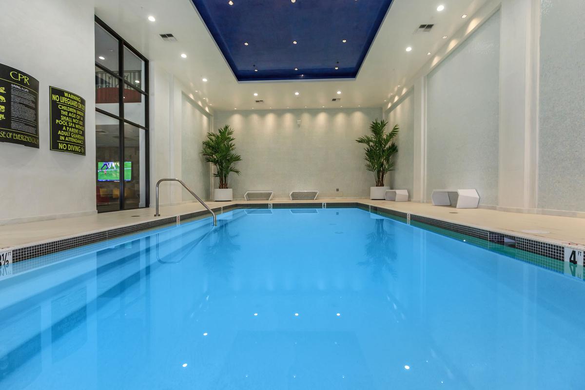 Indoor swimming pool with clear blue water, surrounded by modern lounge chairs and potted plants, featuring a bright ceiling and soft lighting. A large window allows natural light to enter the space.