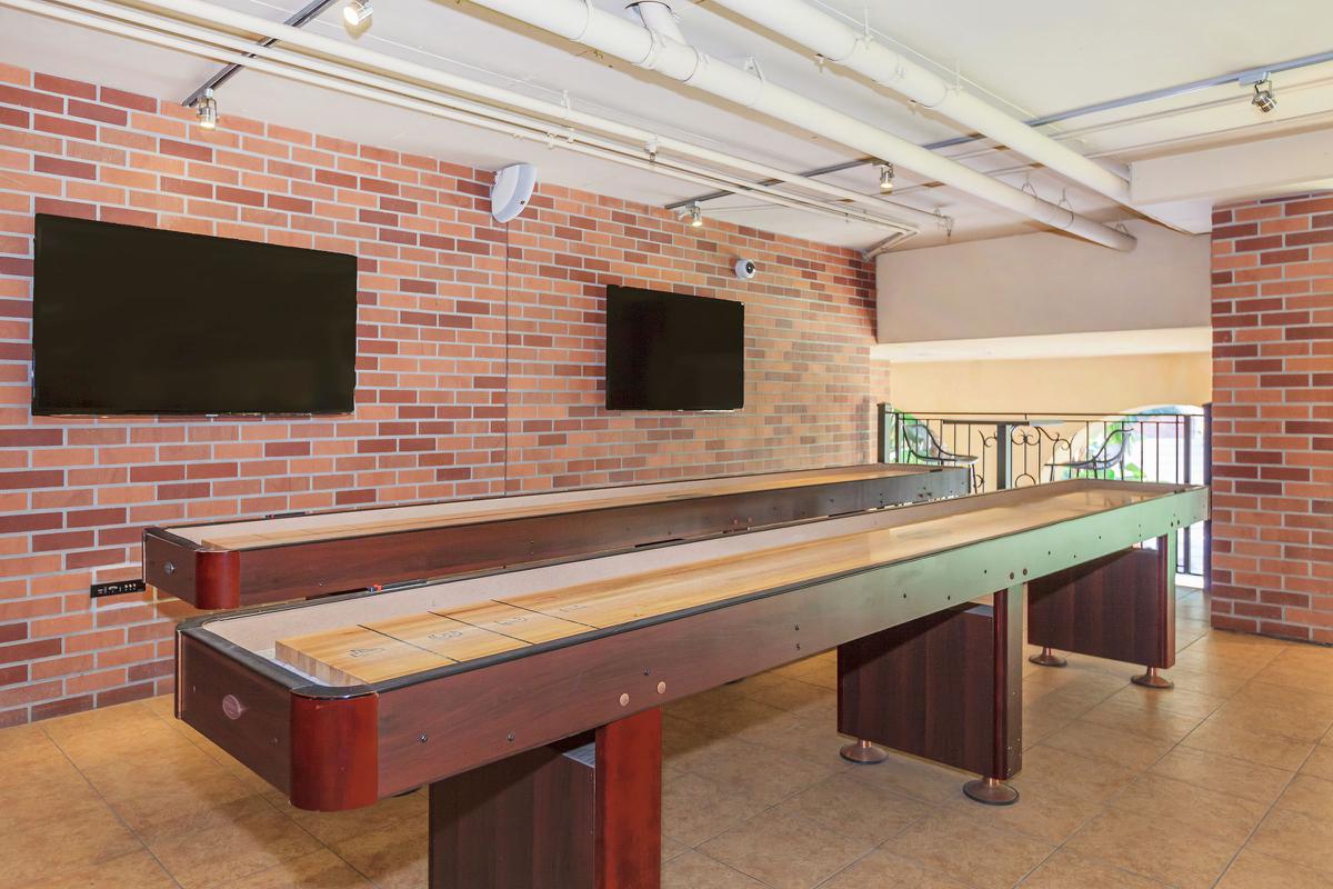 Interior space featuring two shuffleboard tables made of polished wood, set against a backdrop of brick walls. There are two blank television screens mounted on the wall above the tables, and the floor is tiled.