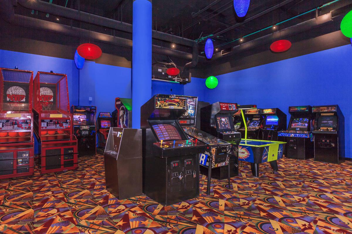 A vibrant arcade room featuring various retro arcade cabinets and games. The walls are painted blue, and colorful pendant lights hang from the ceiling. The flooring has a geometric pattern, enhancing the energetic atmosphere of the space.