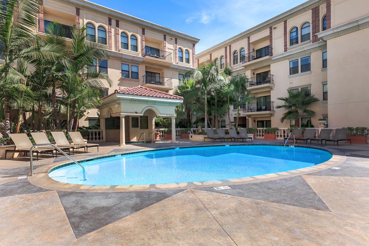 A swimming pool surrounded by lounge chairs and palm trees, with a multi-story building in the background under a clear blue sky.