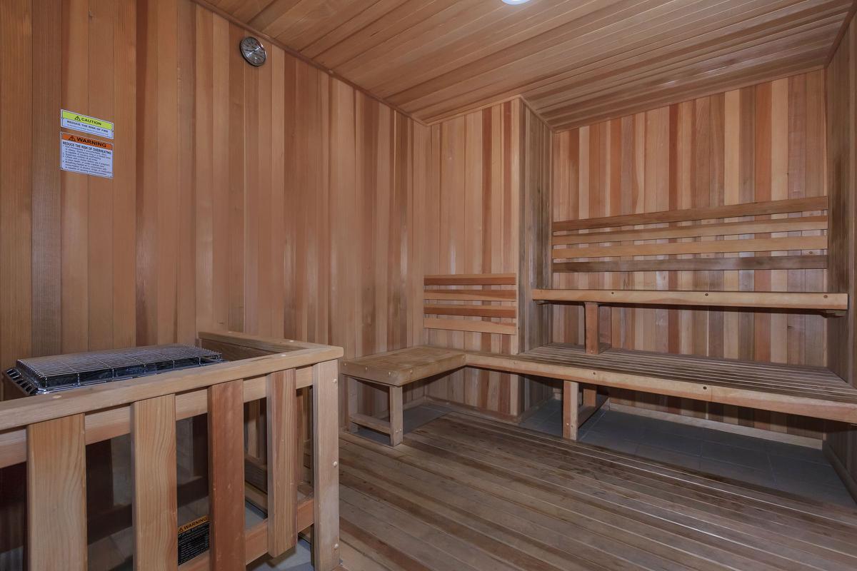 Interior of a wooden sauna featuring wooden benches, a stove, and wood paneled walls. The lighting is soft and warm, creating a relaxing atmosphere.