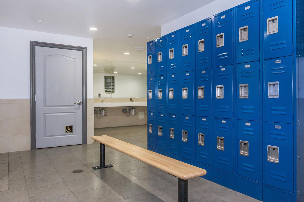 A locker room featuring blue metal lockers along one wall, a wooden bench in front, and a door leading to another area. Mirrors are visible in the background. The floor is tiled and there is a hand dryer mounted on the wall.