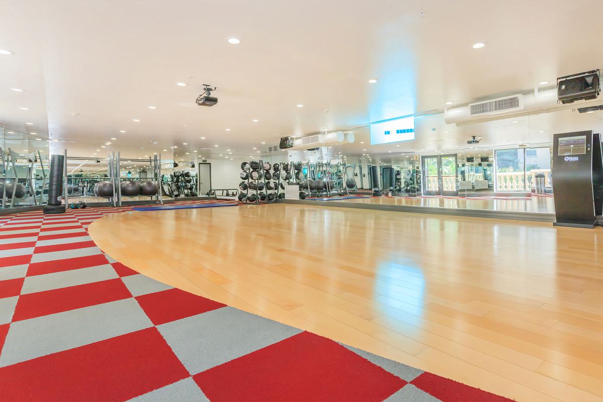 A spacious, well-lit gym featuring wooden flooring with a red and gray patterned area, mirrors on the wall, various workout equipment including weights and exercise balls, and a section for stretching or floor exercises.