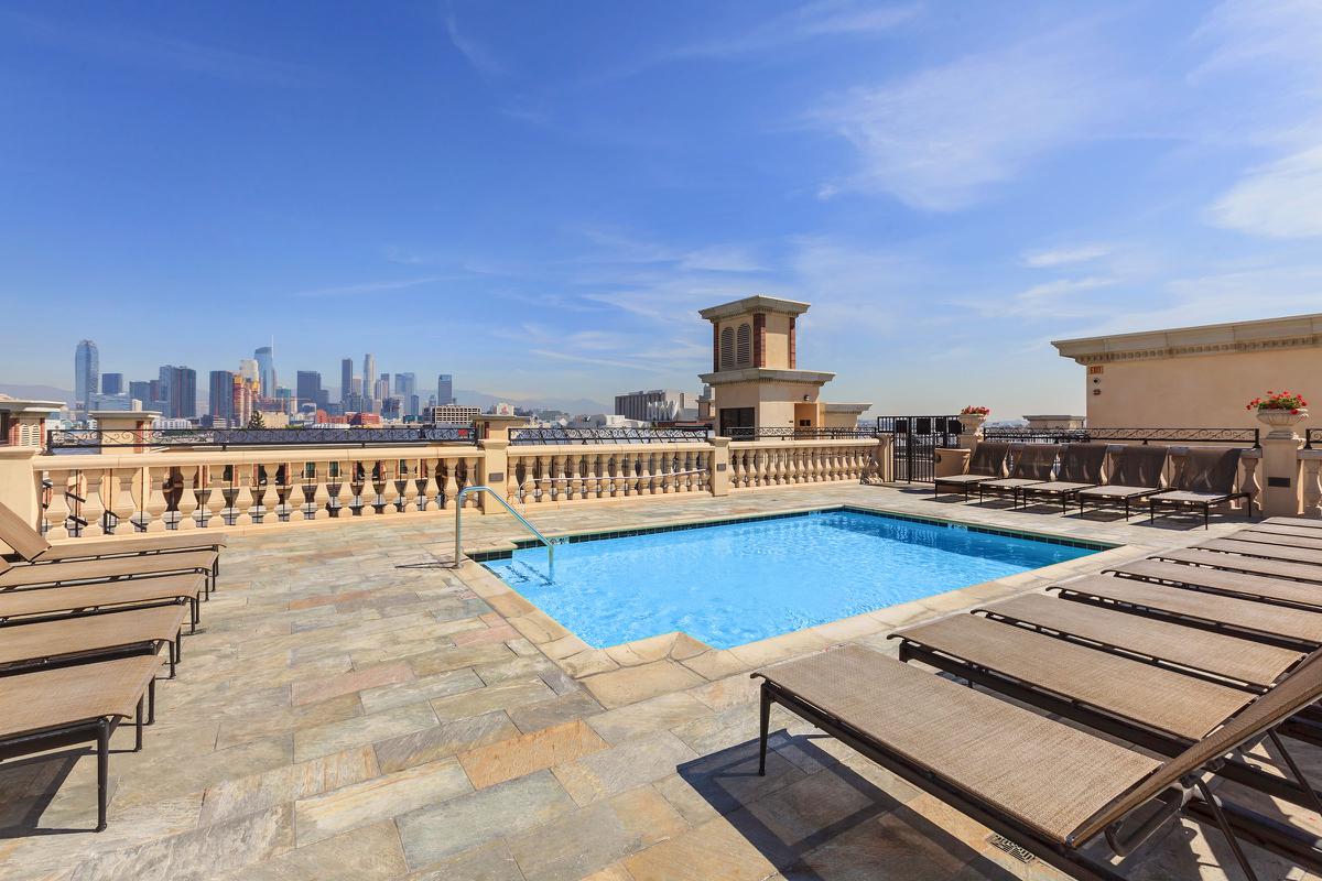 A rooftop pool area with lounge chairs, overlooking a city skyline under a clear blue sky.