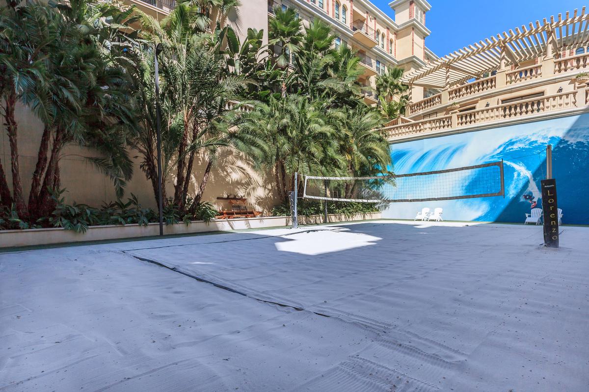 A sandy volleyball court surrounded by lush tropical plants, with a large blue mural depicting a wave on the wall behind. Sunny sky visible above.