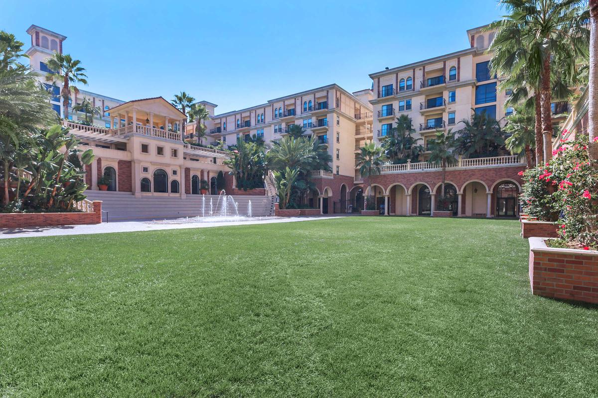 A beautifully landscaped courtyard featuring vibrant green grass, palm trees, and blooming flowers, with a large building in the background that has arches and balconies. A fountain adds a decorative element to the scene, showcasing a sunny and inviting atmosphere.