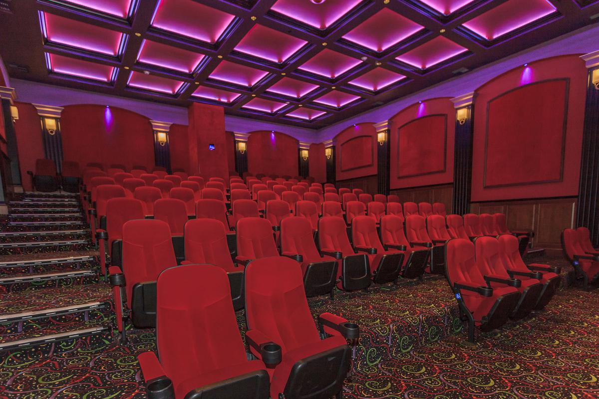 An empty movie theater with red upholstered seats arranged in rows, a colorful patterned carpet, and purple ambient lighting on the coffered ceiling.