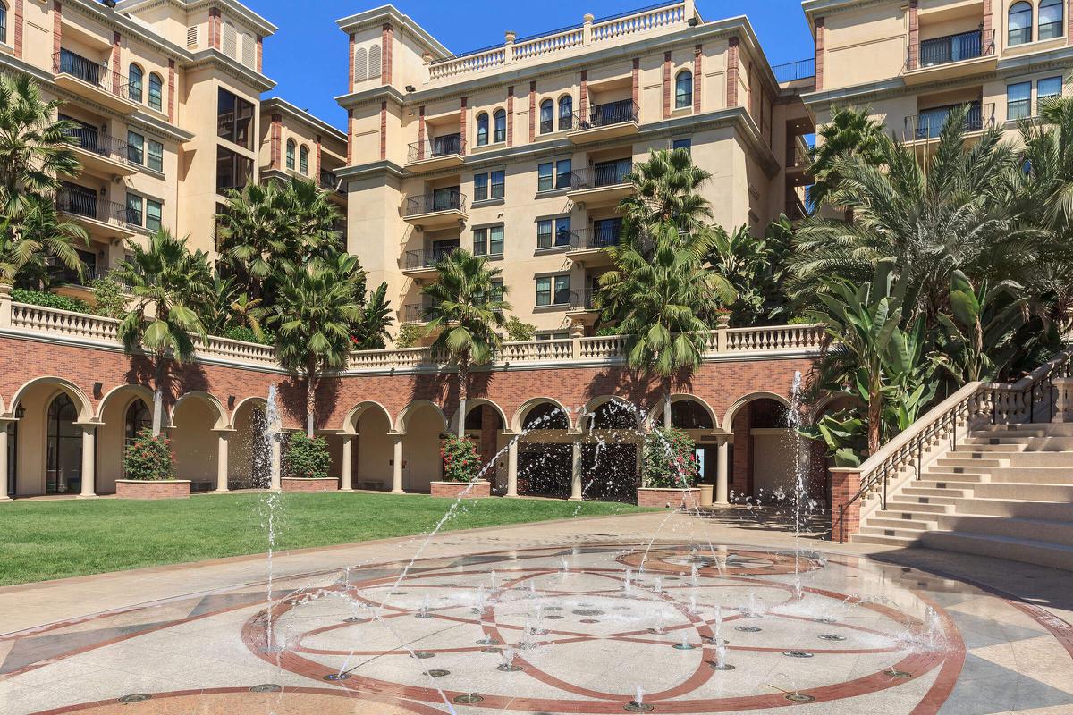 A courtyard featuring a fountain surrounded by lush palm trees and elegant architecture, with a spacious grassy area and a staircase leading to an upper level.