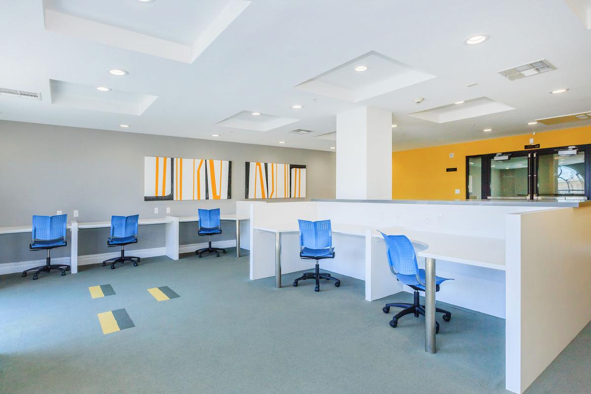 An interior space with a modern design featuring several empty desks with blue chairs, a green carpet, and abstract wall art. The walls are painted in gray and yellow tones, with ample lighting from the ceiling.