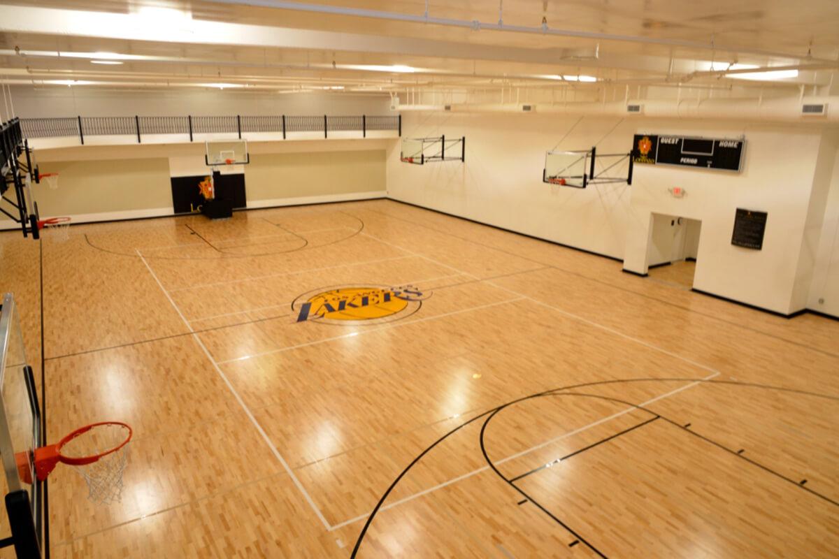 An indoor basketball court featuring a wood floor, basketball hoops at both ends, and the logo of the Los Angeles Lakers at the center. The court is well-lit and spacious, with a black railing visible in the background.