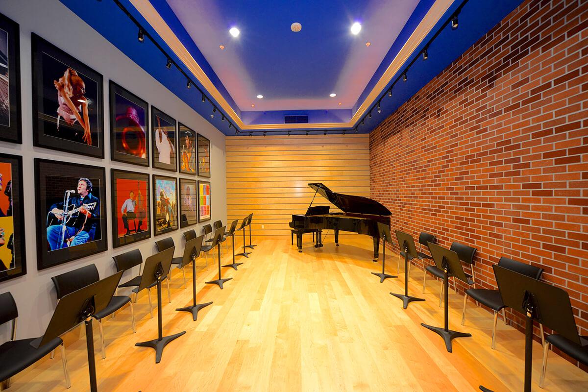 A spacious music room with a grand piano at the center, surrounded by rows of black chairs and adorned with framed music-related artwork on the walls. The room features a wood floor and a ceiling with blue accents, creating a bright and inviting atmosphere.