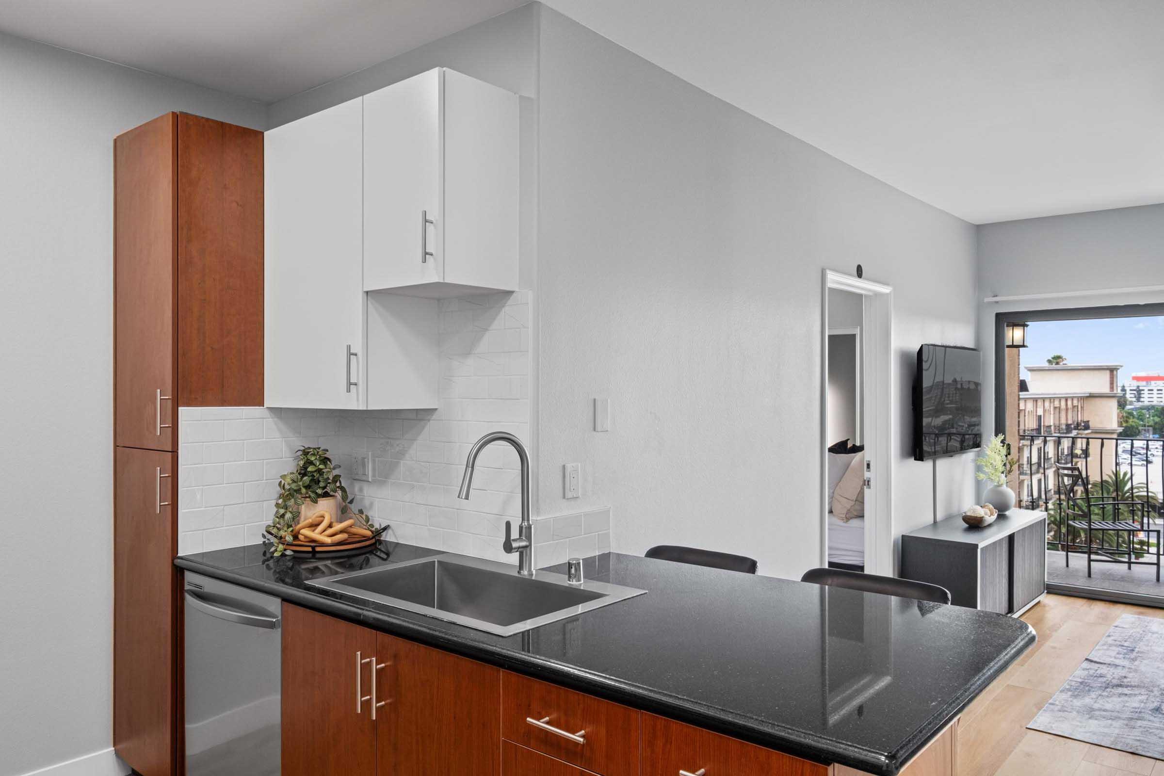 Modern kitchen featuring a sleek countertop with a stainless steel sink, wooden cabinetry, and a white backsplash. A bowl of bananas is displayed on the counter. The background shows a living area with a television and a glimpse of outdoor space through sliding doors.
