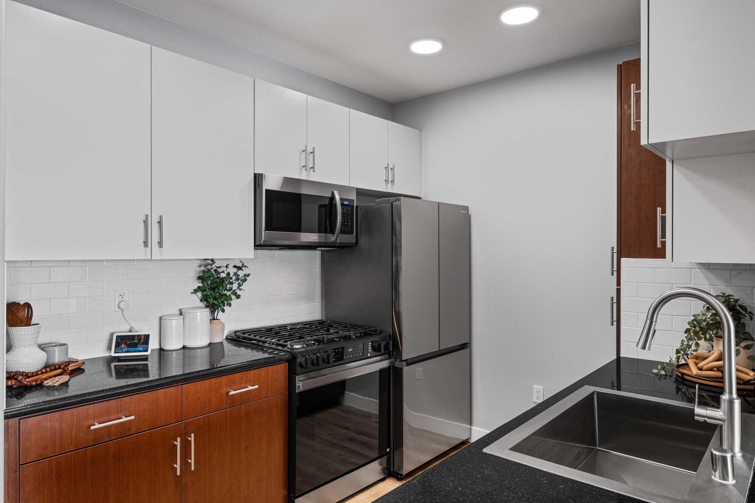 A modern kitchen featuring black countertops, stainless steel appliances including a microwave and refrigerator, white cabinets, and a wood-paneled cabinet. A sink is visible, along with a decorative plant and kitchen essentials, creating a clean and stylish cooking space.