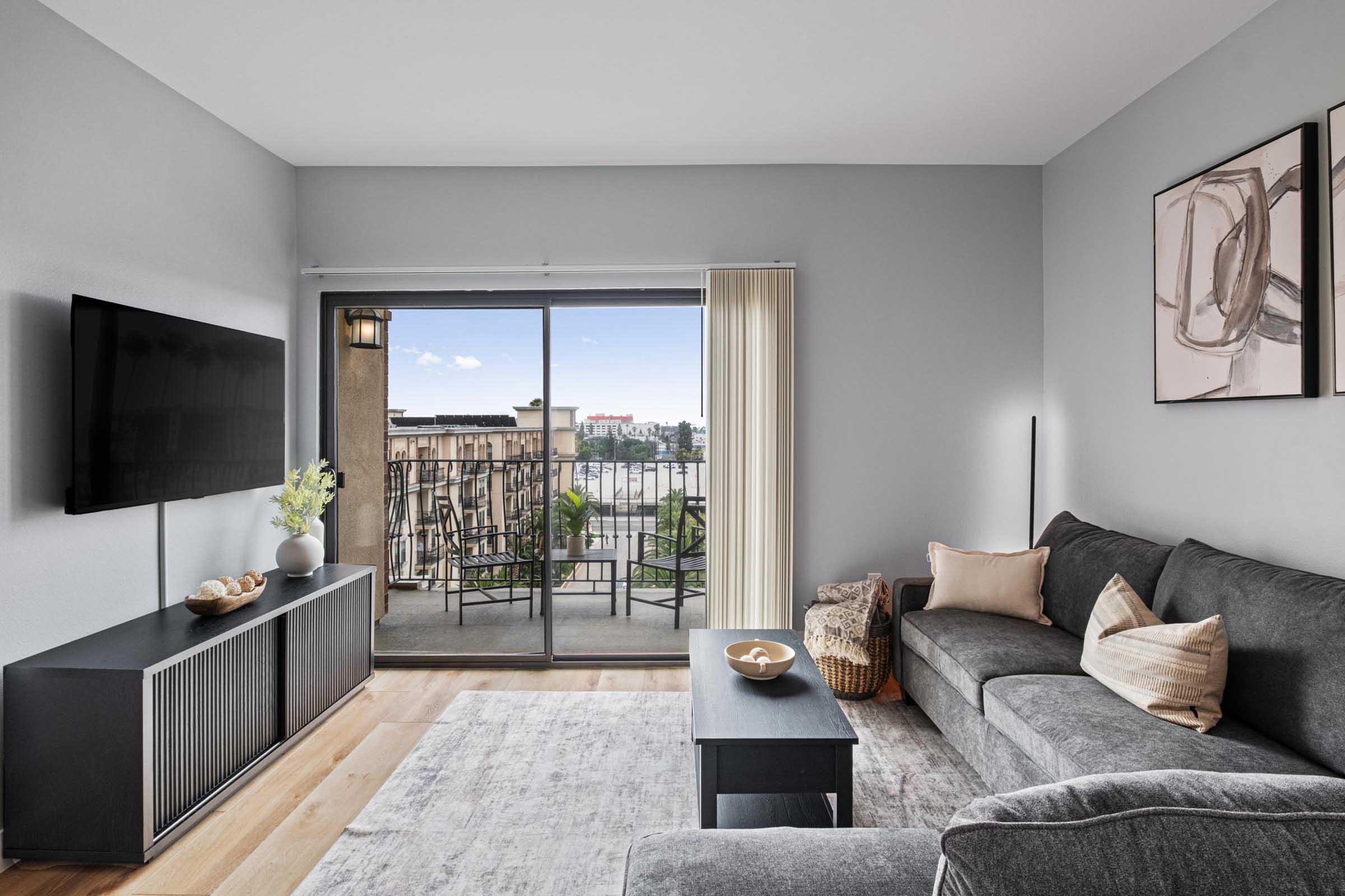 A modern living room featuring a gray sofa, a black coffee table, and a large wall-mounted TV. A sliding glass door leads to a balcony with outdoor seating, allowing natural light to fill the space. Decorative elements include a bowl of fruit and artwork on the walls, creating a stylish and inviting atmosphere.