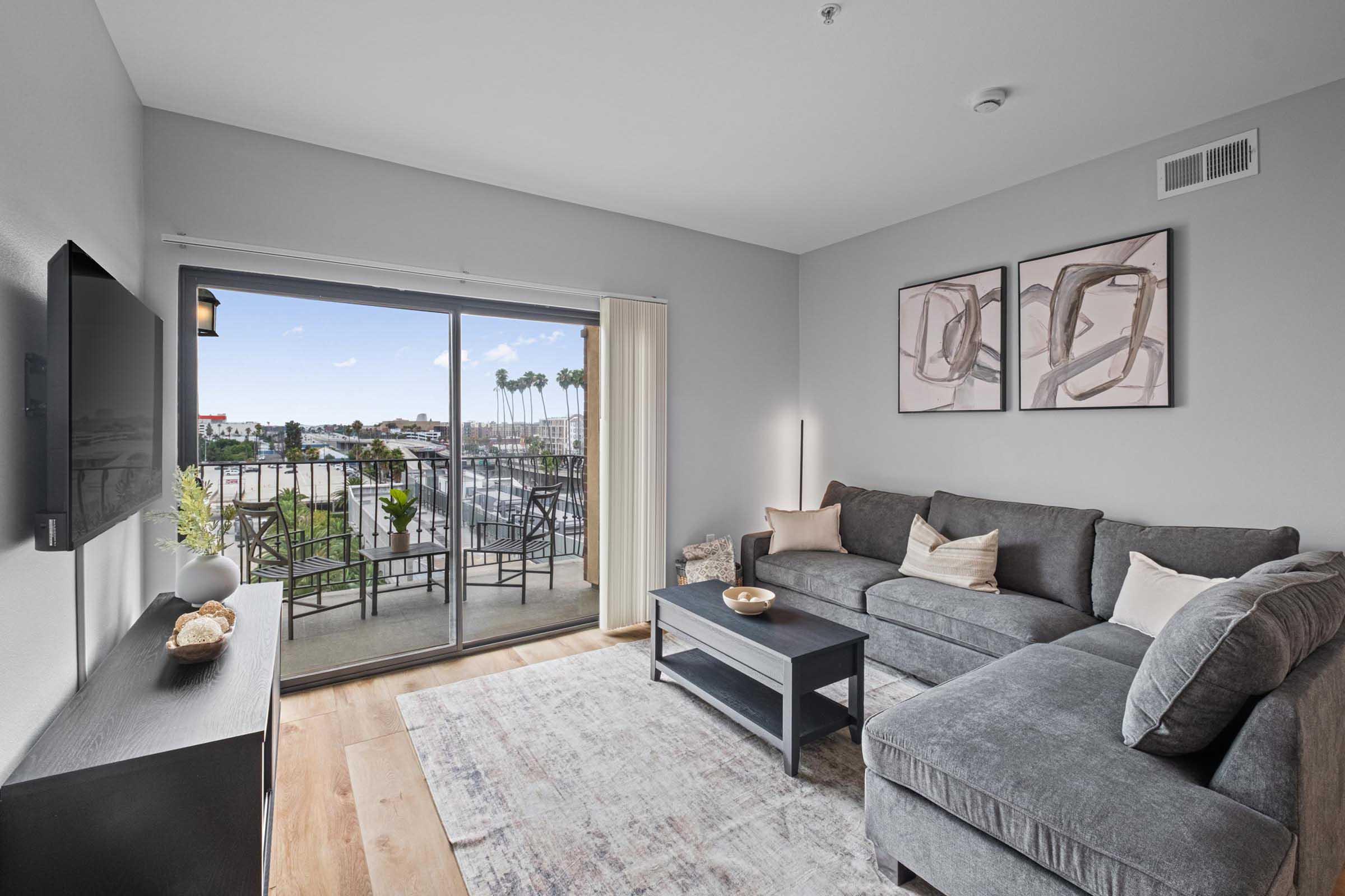 A modern living room featuring a grey sectional sofa, a black coffee table, and a wall-mounted TV. Large sliding glass doors lead to a balcony with a view of palm trees. Light walls and a textured rug create a cozy atmosphere, complemented by framed artwork on the wall.