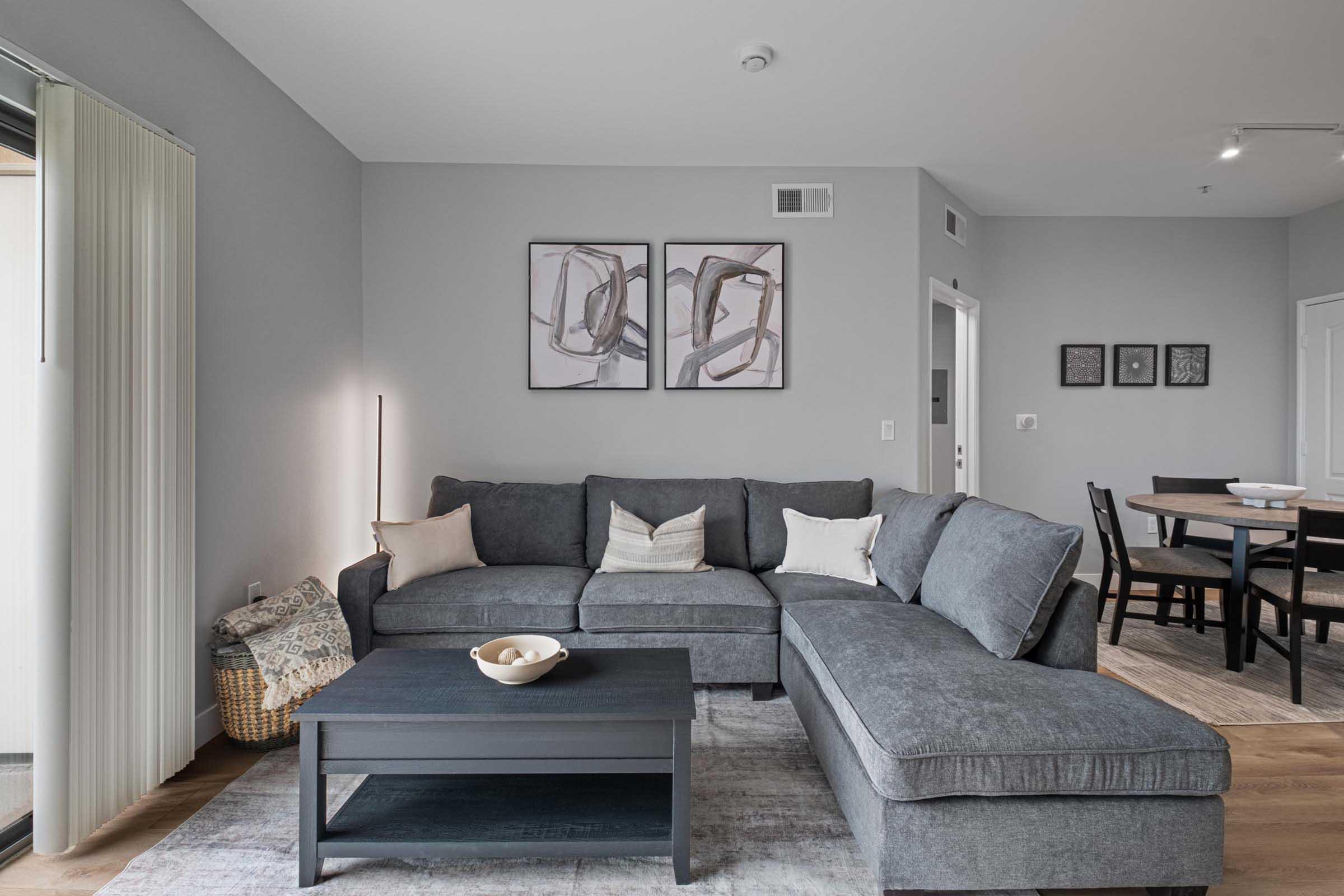 A modern living room featuring a gray sectional sofa adorned with decorative pillows, a dark coffee table, and a bowl of decorative items on top. Two abstract framed artworks hang on the wall. Natural light filters through sheer curtains, and a dining area with a table and chairs is visible in the background.