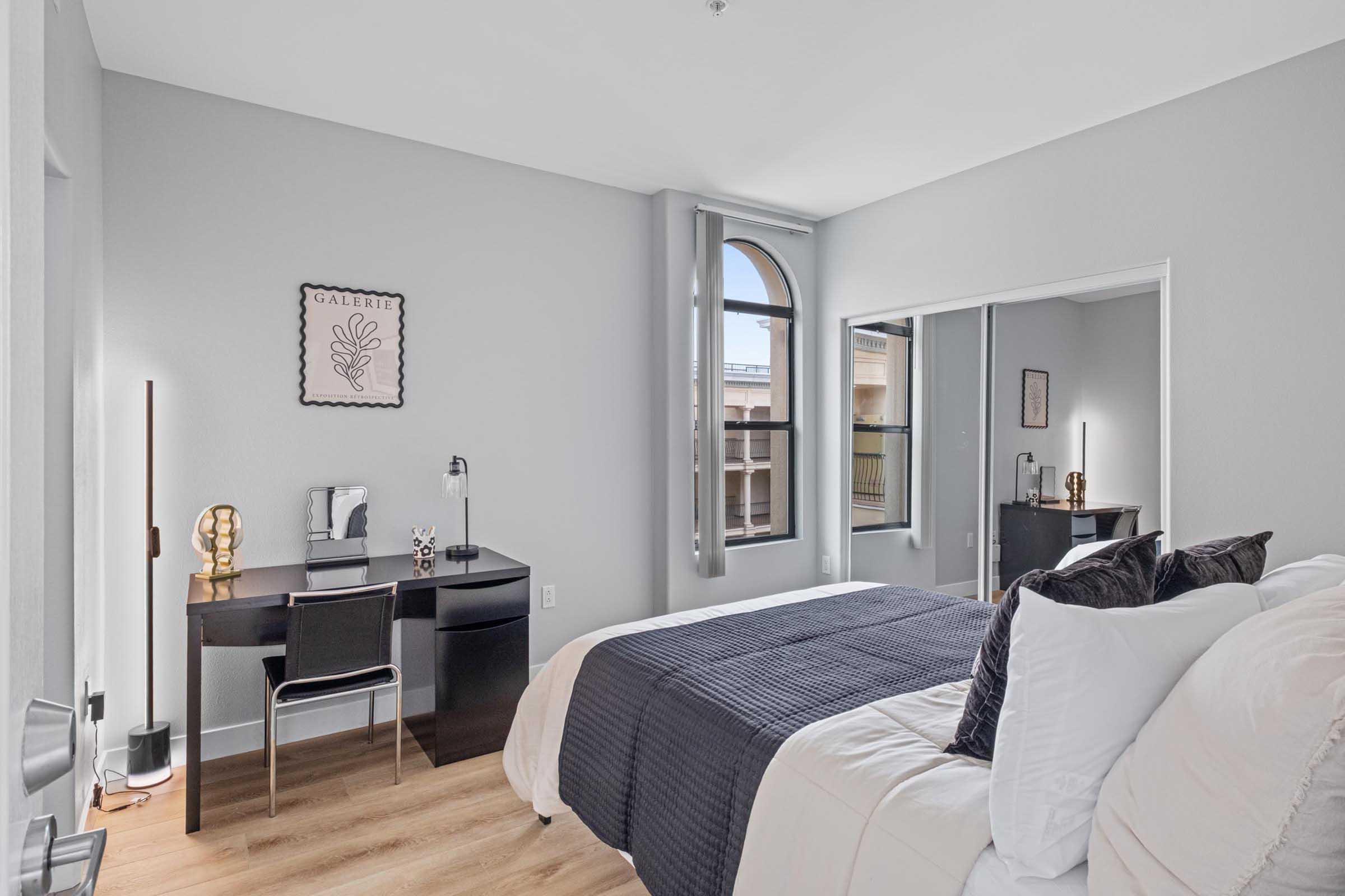 Bright and modern bedroom featuring a neatly made bed with white and navy bedding, a stylish desk with a lamp and chair, and a mirror. The room has large windows letting in natural light, grey walls, and minimalistic decor, creating a clean and inviting atmosphere.