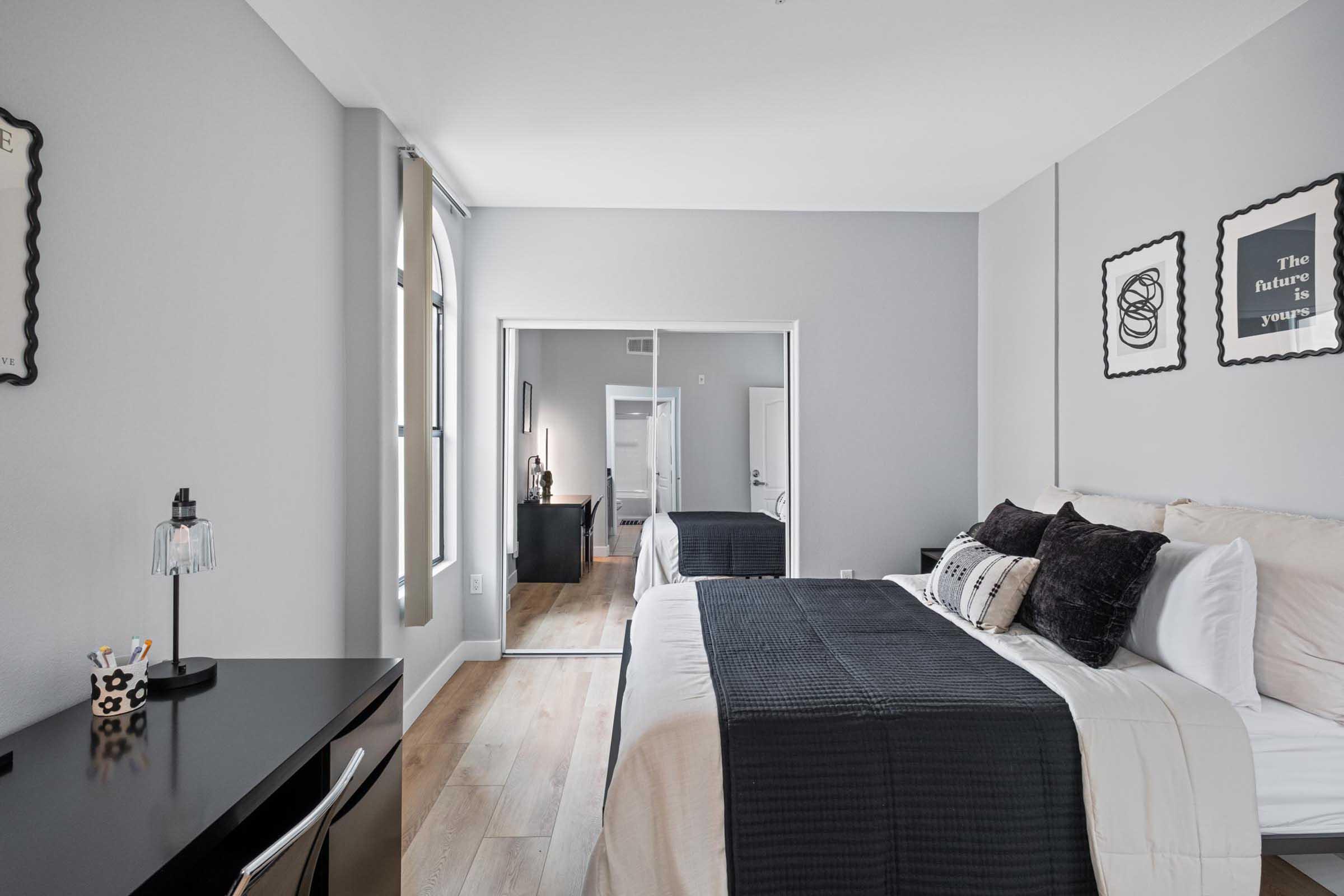 A stylish bedroom featuring a large bed with black and white bedding, a minimalist desk with stationery, and a full-length mirror. The room has light gray walls and wooden flooring, creating a modern and serene atmosphere. Decorative art pieces adorn the walls, enhancing the contemporary design.