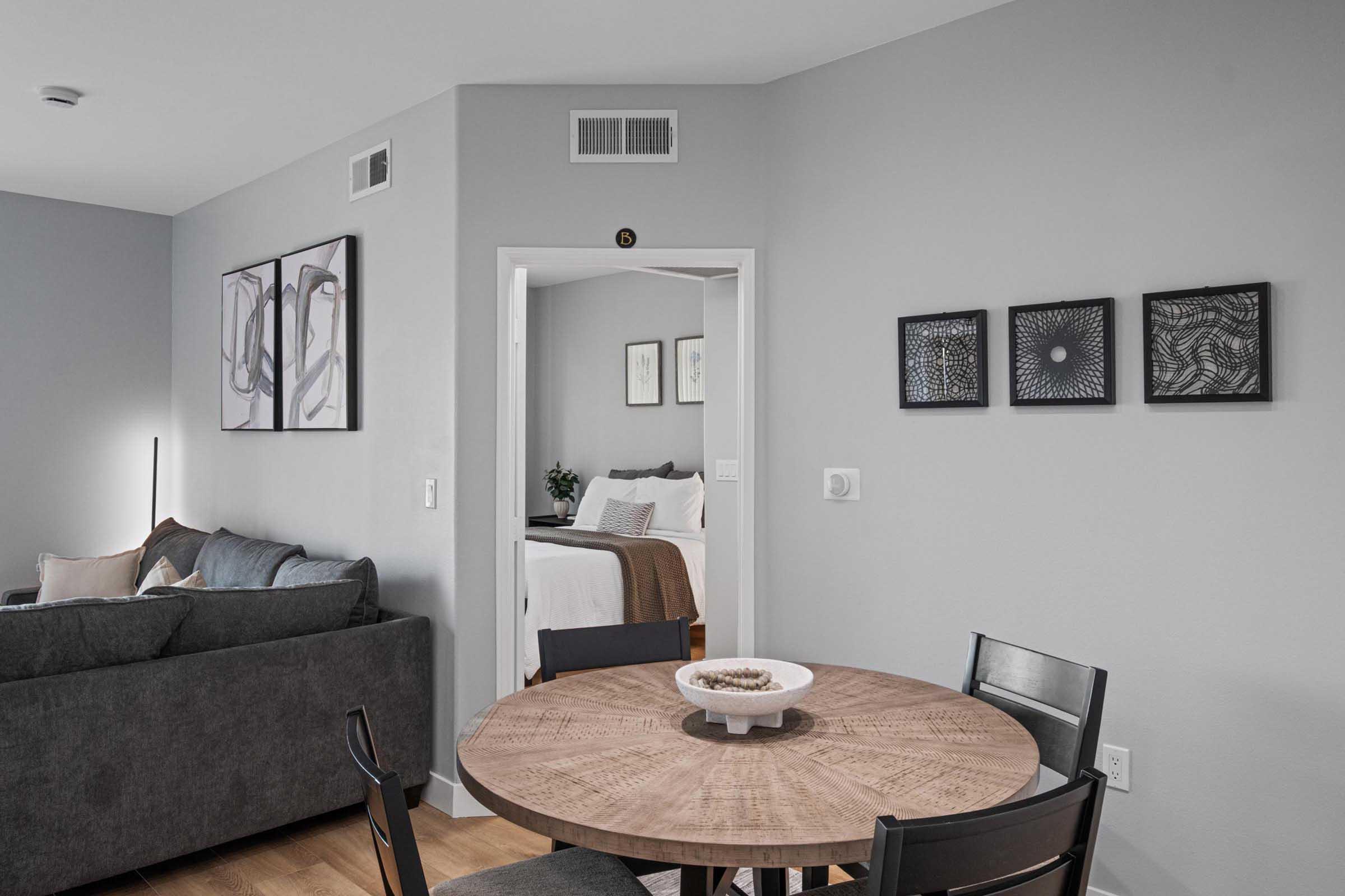 A cozy, modern living space featuring a round wooden dining table with black chairs. In the background, a gray sofa and wall art adorn the walls. A doorway leads to a bedroom, and light streams in through the room, creating a welcoming atmosphere.