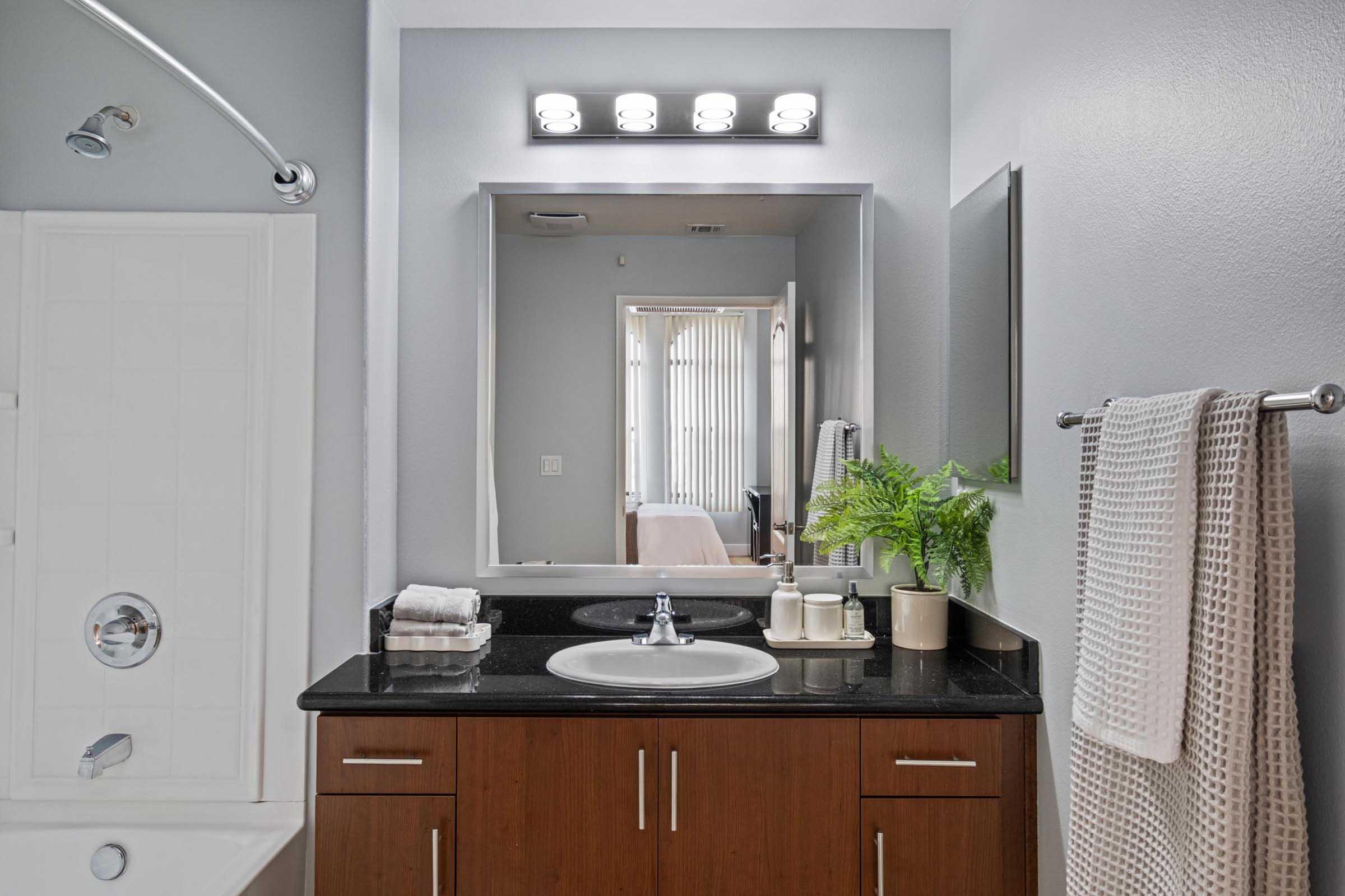 A clean and modern bathroom featuring a black countertop with a sink, neatly arranged toiletries, and a large mirror above. A light fixture with multiple bulbs illuminates the space, while a shower with a glass door is visible on the left. A plant adds a touch of greenery, and a towel hangs on a rack nearby.
