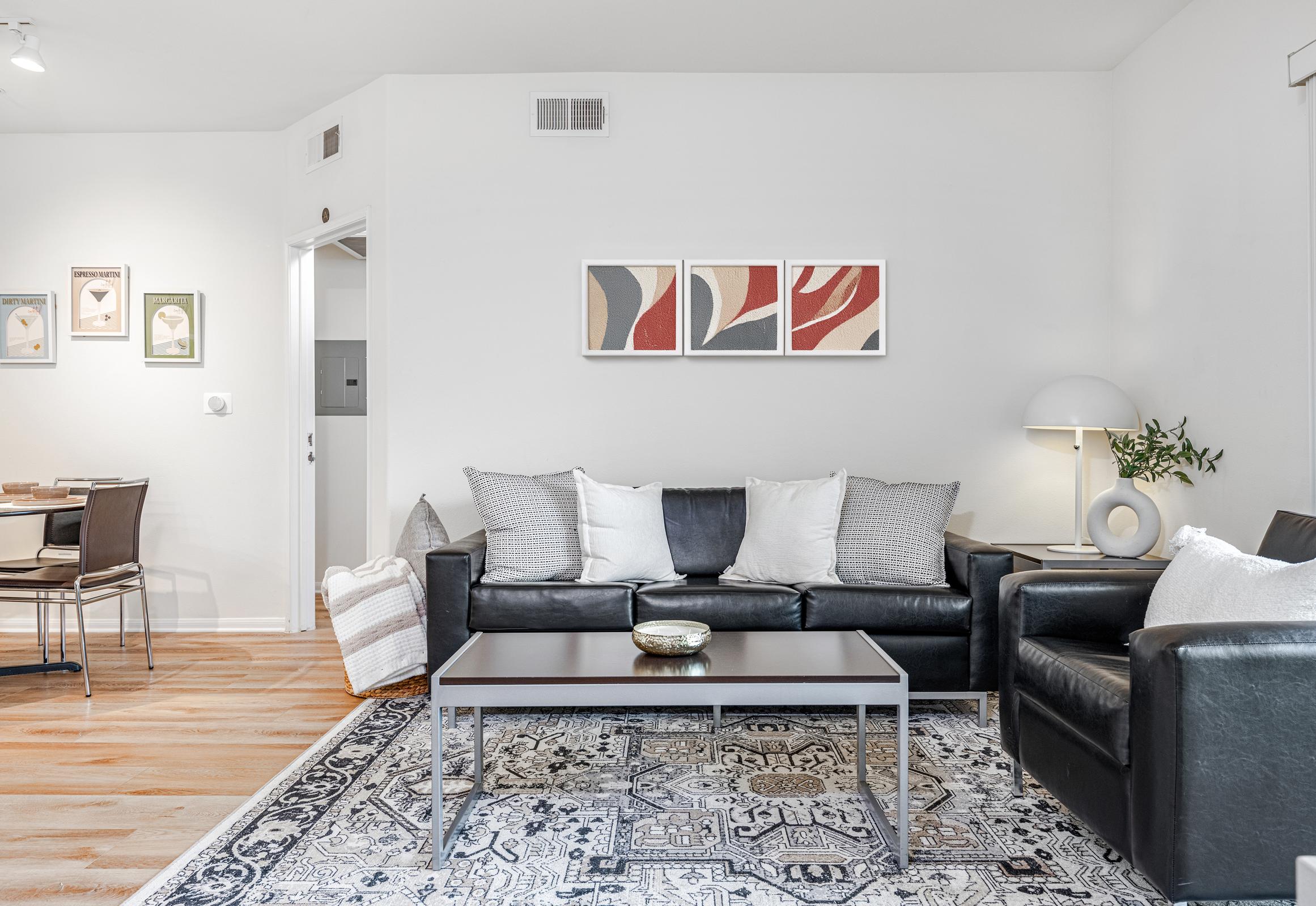 A modern living room featuring a black leather sofa with decorative pillows, a matching armchair, and a coffee table. The space is adorned with a patterned rug and wall art consisting of abstract designs. A lamp and a small plant add a touch of elegance to the inviting atmosphere.