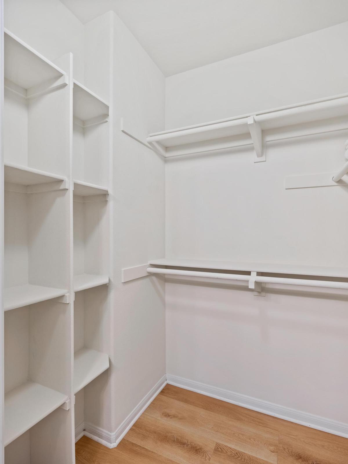 Empty walk-in closet featuring white shelving units on the left side and hanging rods along the back wall. The floor is finished with light brown wood, and the walls are painted white, creating a bright and clean storage space.