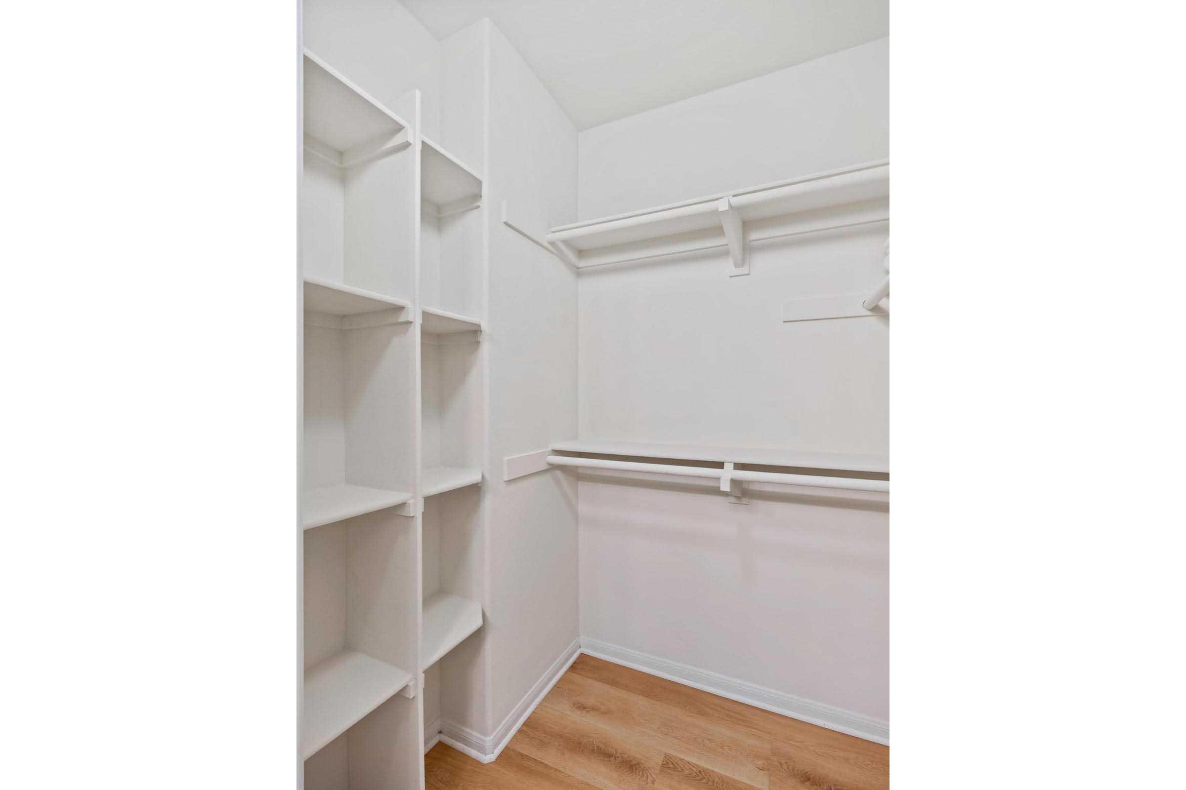 An empty walk-in closet featuring white shelves and hanging rods against plain white walls. The floor is wooden, and the overall space is well-lit and spacious, emphasizing its potential for organization and storage.