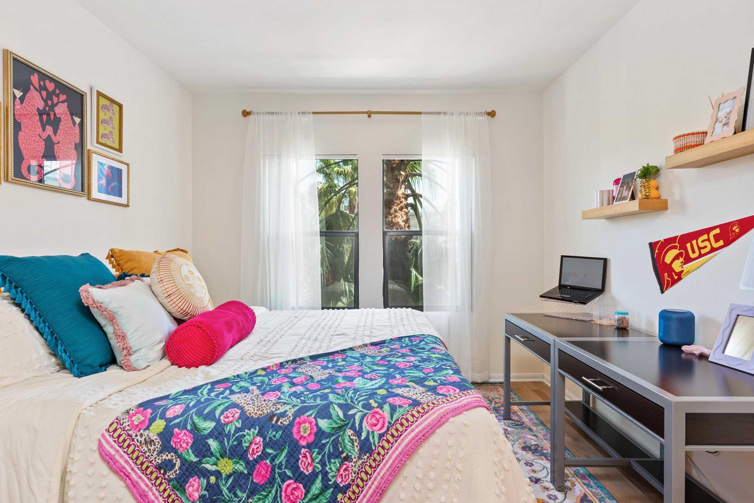 A cozy bedroom featuring a bed with colorful pillows and a decorative quilt. There is a small desk with a laptop, and decorative wall art is displayed. Large windows with sheer curtains allow natural light to fill the room, showcasing palm trees outside. The overall decor is vibrant and inviting.