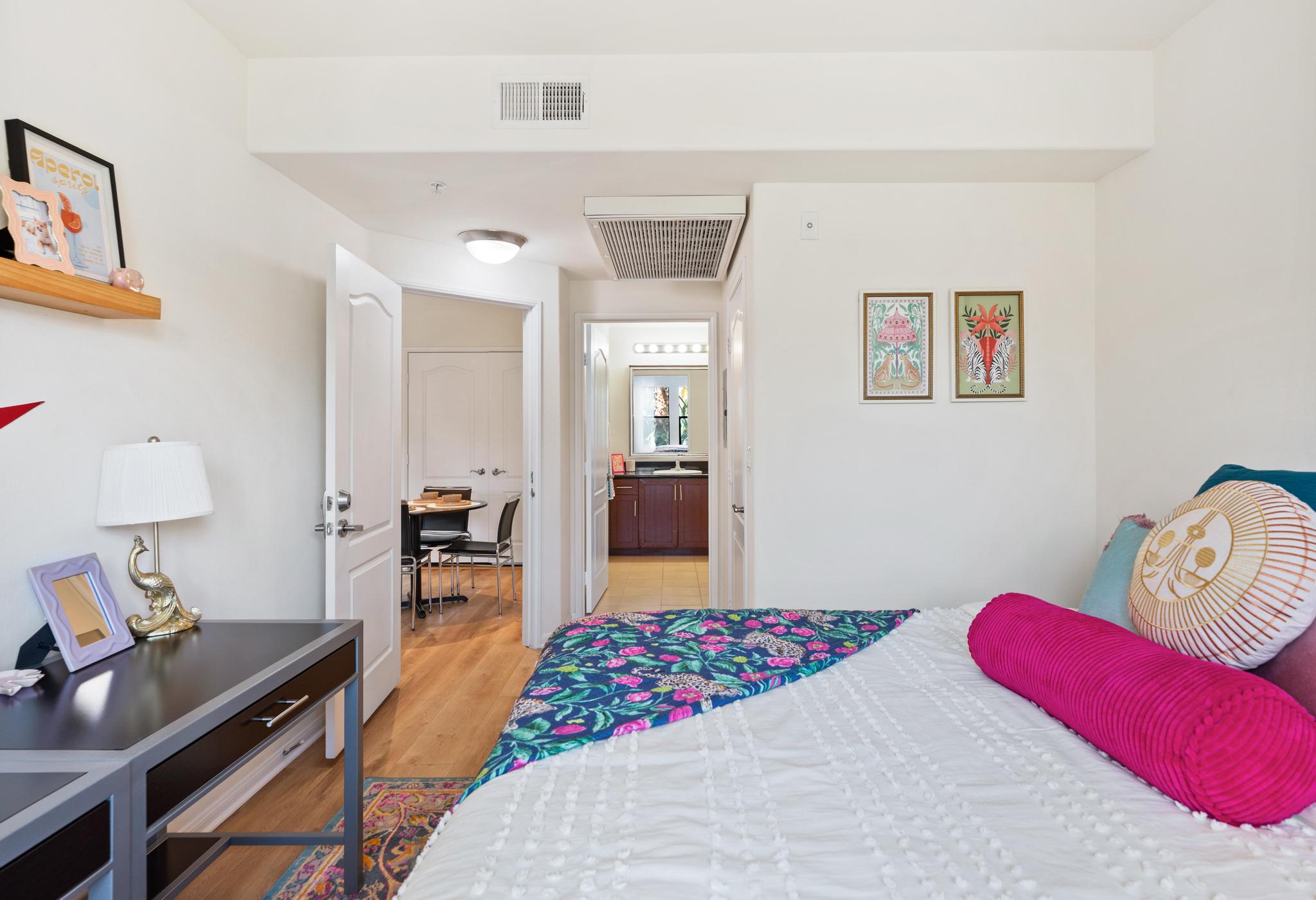 A bright, modern bedroom featuring a bed with a colorful decorative pillow, a nightstand with a lamp, and framed artwork on the walls. A doorway leads to a hallway with a kitchen visible in the background. Natural light fills the space, enhancing the warm and inviting atmosphere.