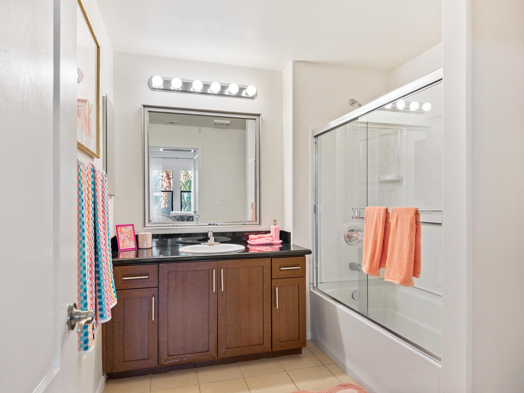 A bright bathroom featuring a dark wood vanity with a sink and a large mirror above. The space includes a bathtub with a glass shower door, light-colored tiles, and colorful towels hanging on a rack. Natural light enters through a window, enhancing the airy feel of the room.