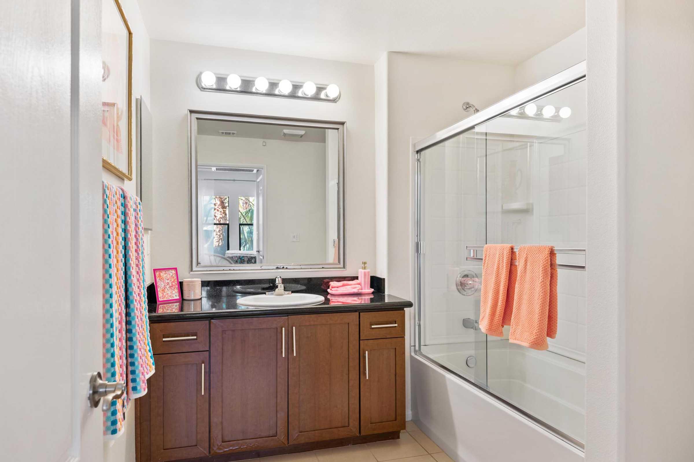 A modern, well-lit bathroom featuring a double sink vanity with dark wood cabinetry, a large mirror, and overhead lighting. The space includes a glass-enclosed shower and a bathtub. Two colorful towels hang on a towel rack, and there's a window allowing natural light. The decor is clean and contemporary.