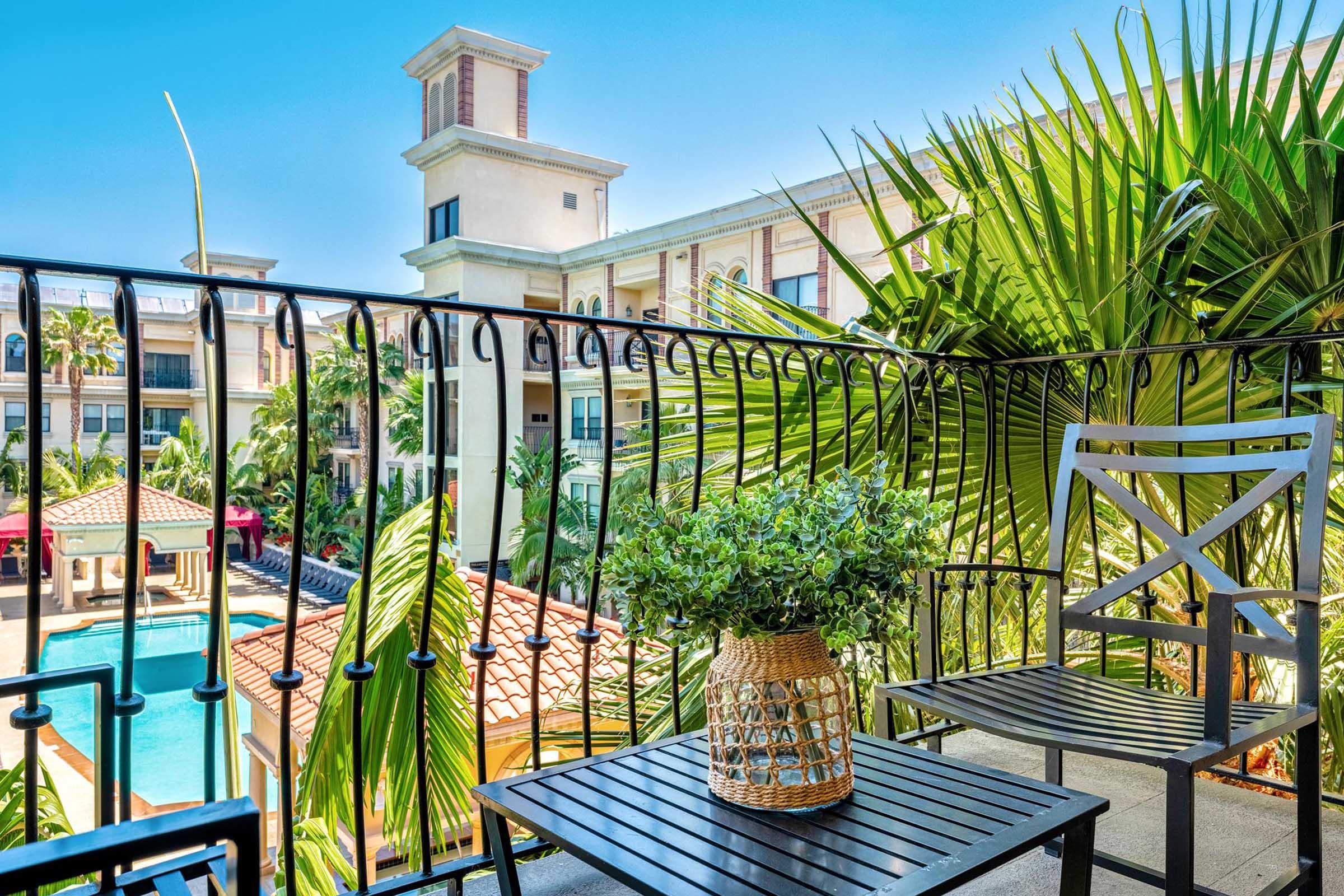 A balcony view featuring a potted plant on a small black table and a chair, overlooking a swimming pool surrounded by palm trees and tropical foliage. In the background, a building with a light-colored façade and multiple balconies can be seen, all under a clear blue sky.