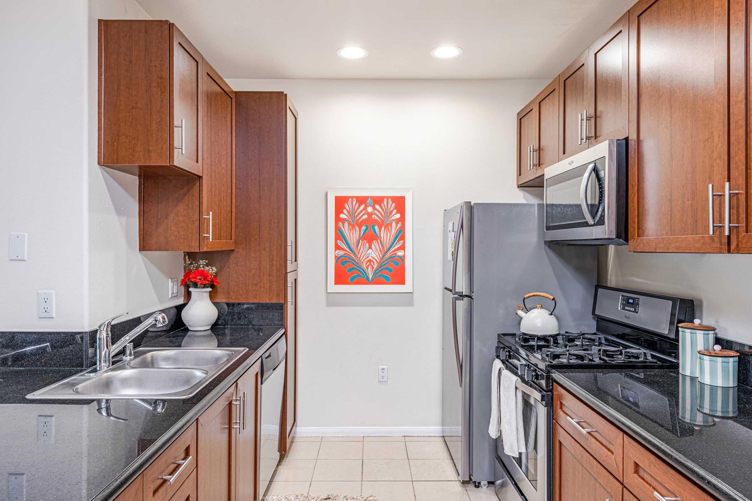 A modern kitchen featuring wooden cabinetry, a stainless steel refrigerator, and an oven/stovetop. The countertops are black granite. A white vase with red flowers sits on the counter, and a colorful artwork with floral design is displayed on the wall. Bright lighting enhances the inviting atmosphere.