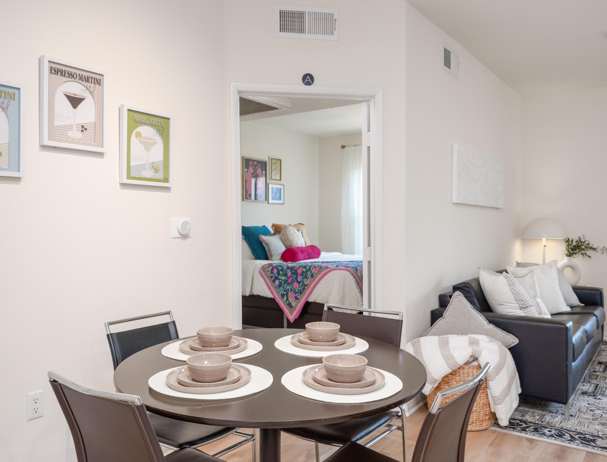 A modern living space featuring a round dining table set for four with brown plates and placemats. A cozy couch is in the foreground, along with decorative pillows and a basket. In the background, a doorway leads to a bedroom with a bed and artwork on the walls, creating a warm, inviting atmosphere.