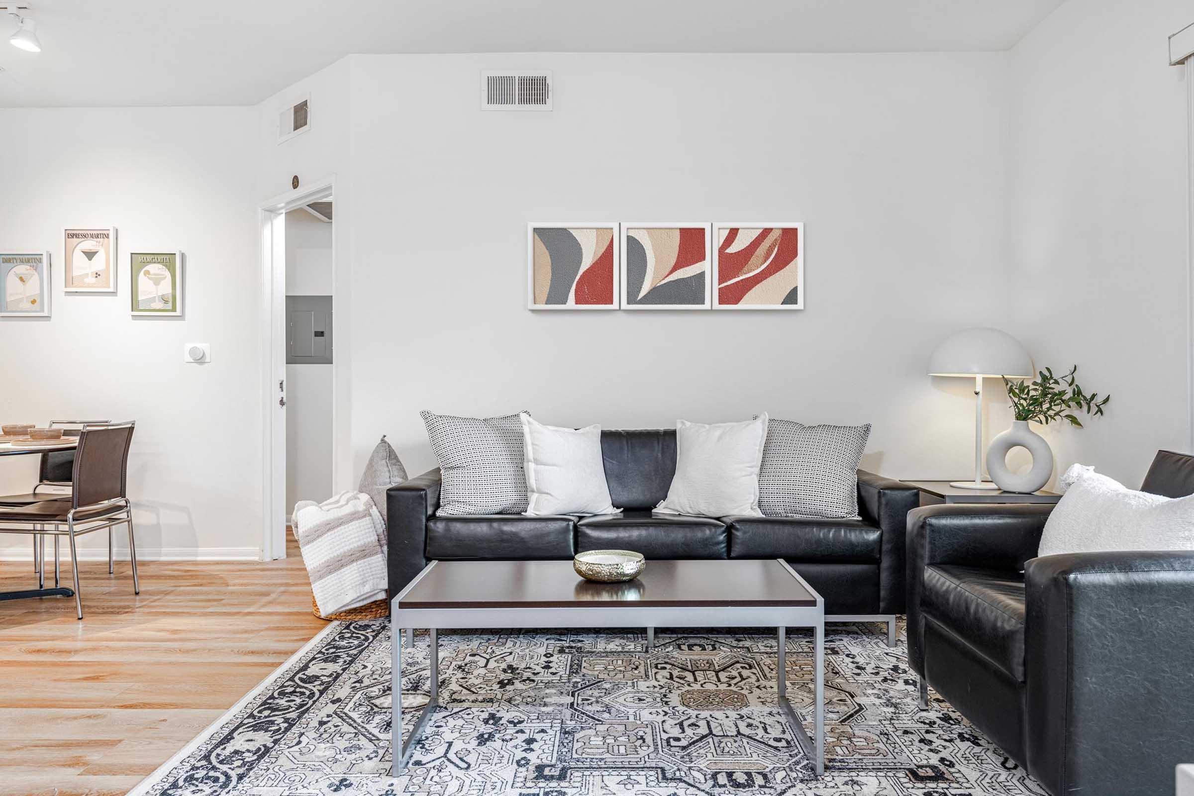 A modern living room featuring a black leather sofa and armchair adorned with decorative pillows. A coffee table sits in the center on a patterned rug. On the wall, three abstract art pieces are displayed. A stylish lamp and a dining area with a round table and chairs are visible in the background.
