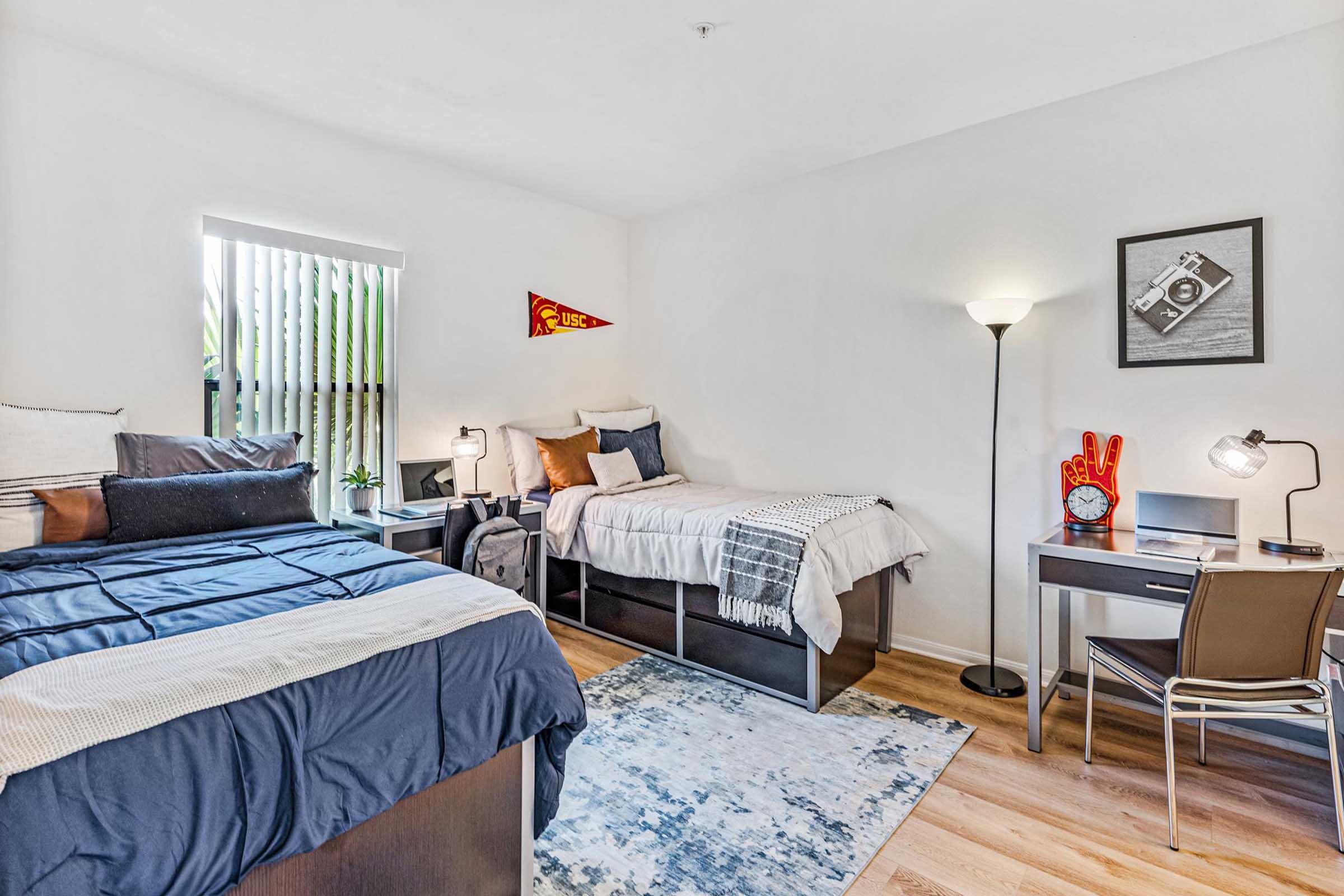 A cozy bedroom with two beds—one with blue bedding and the other with neutral tones. A desk with a chair is positioned against the wall, accompanied by a lamp and a small plant. A USC pennant hangs on the wall, and natural light filters in through a window with wooden blinds. A patterned rug adds warmth to the space.